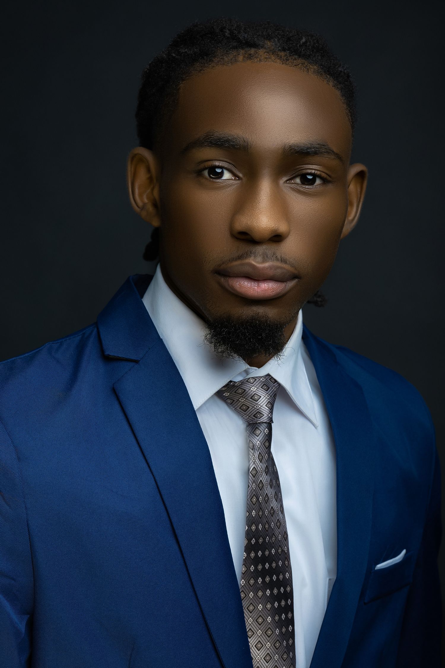 Professional headshot of a businessman wearing a navy blue suit and polka dot tie against a dark background.