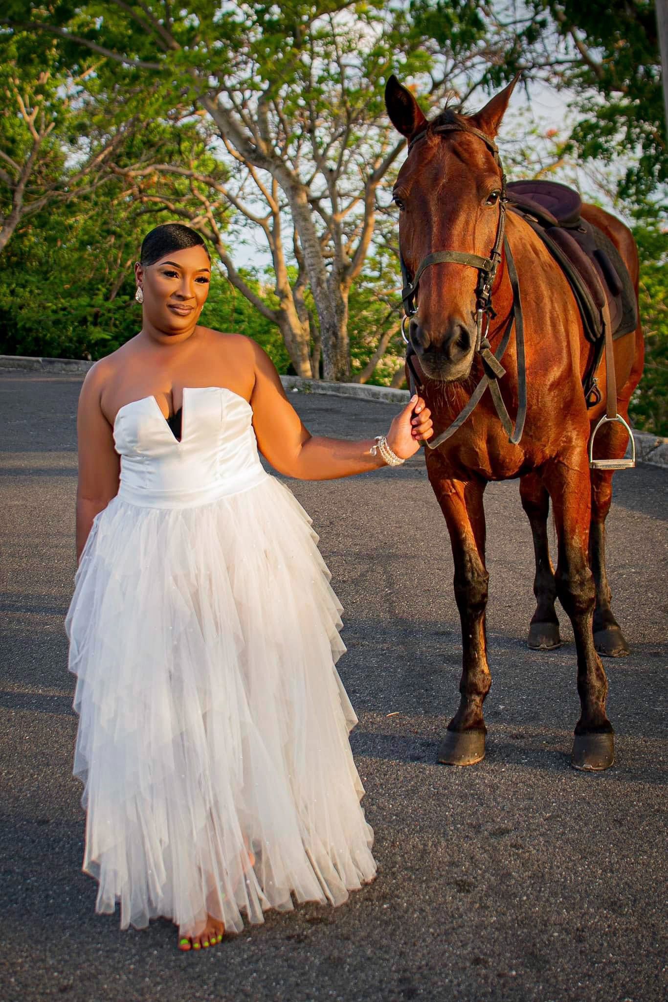Bride in strapless tulle gown poses with brown horse on paved path.