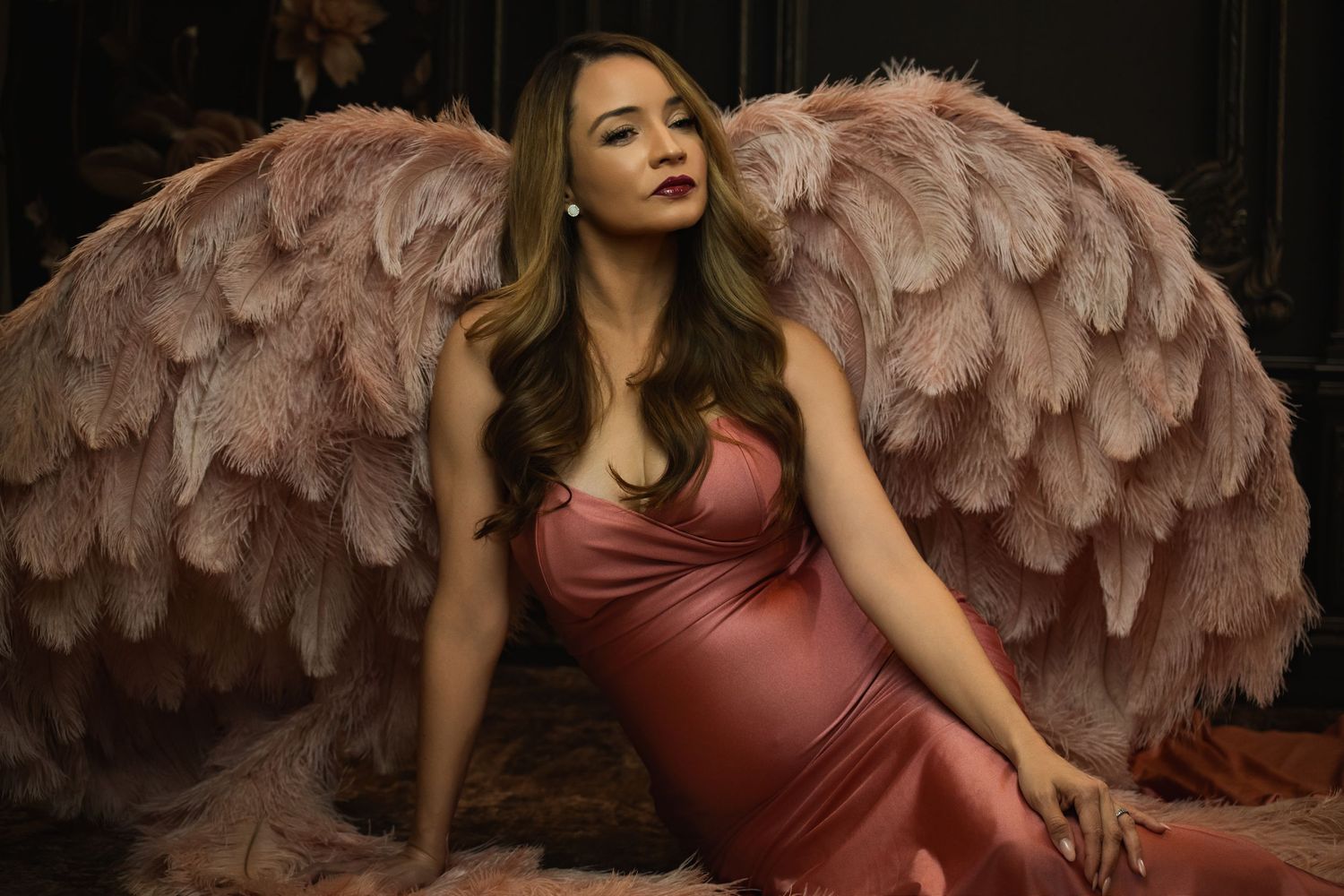 Maternity photo of woman in silk dress with large feathered angel wings against dark background.