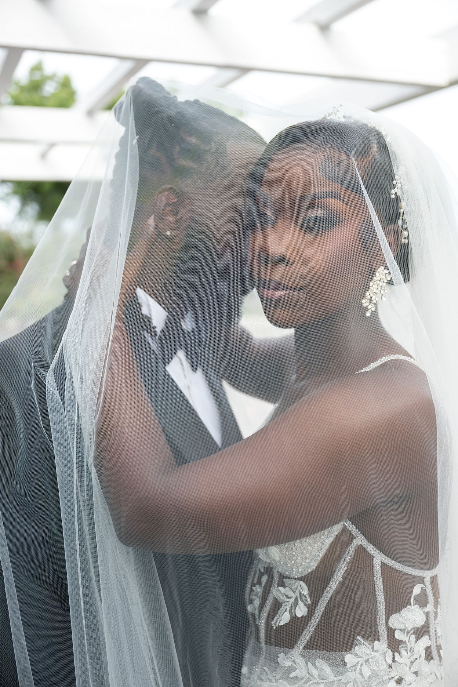 Bride and groom share an intimate moment under a white wedding veil, bride wearing lace gown and crystal earrings.