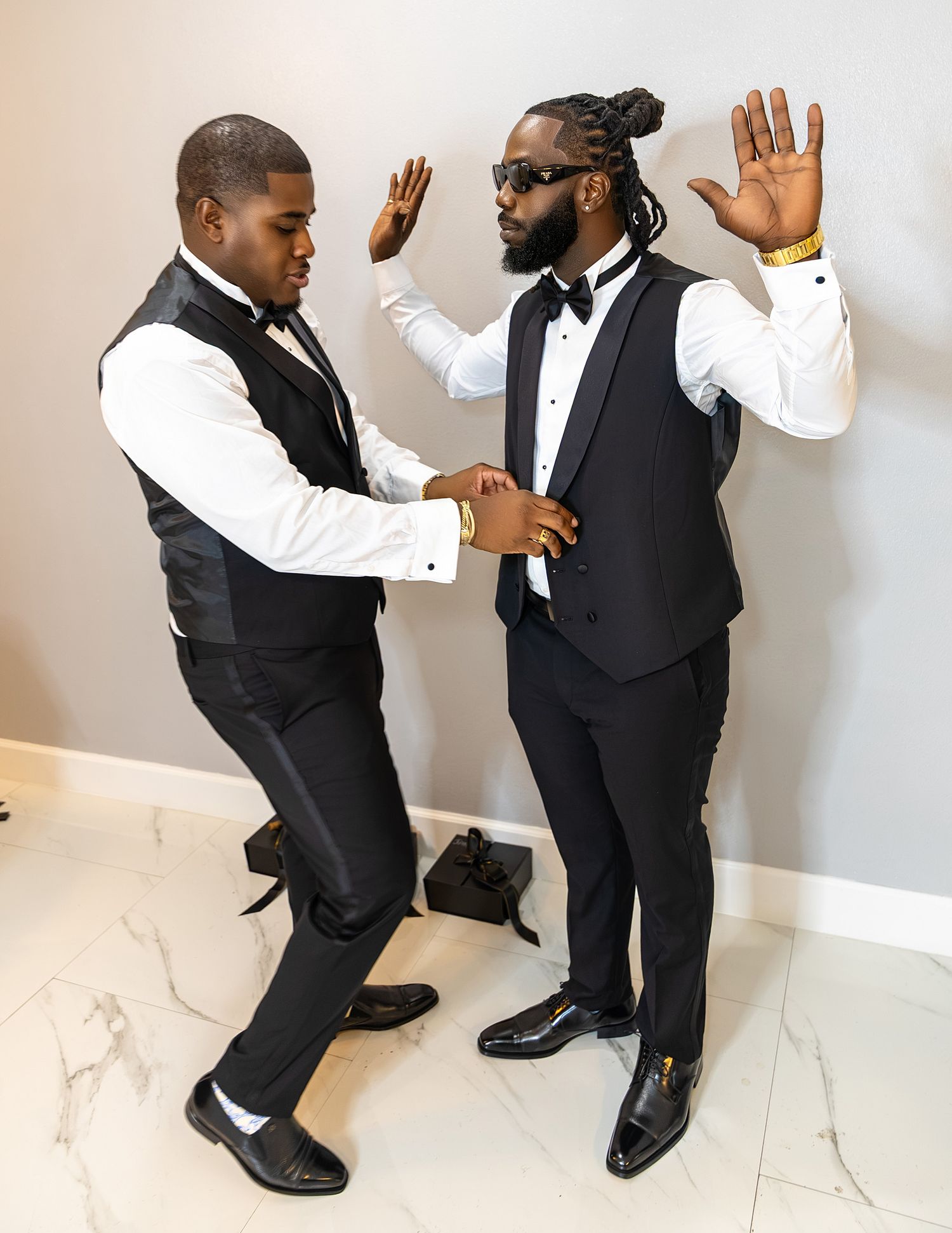 Two men in formal black tuxedos with bow ties pose playfully indoors on marble flooring.