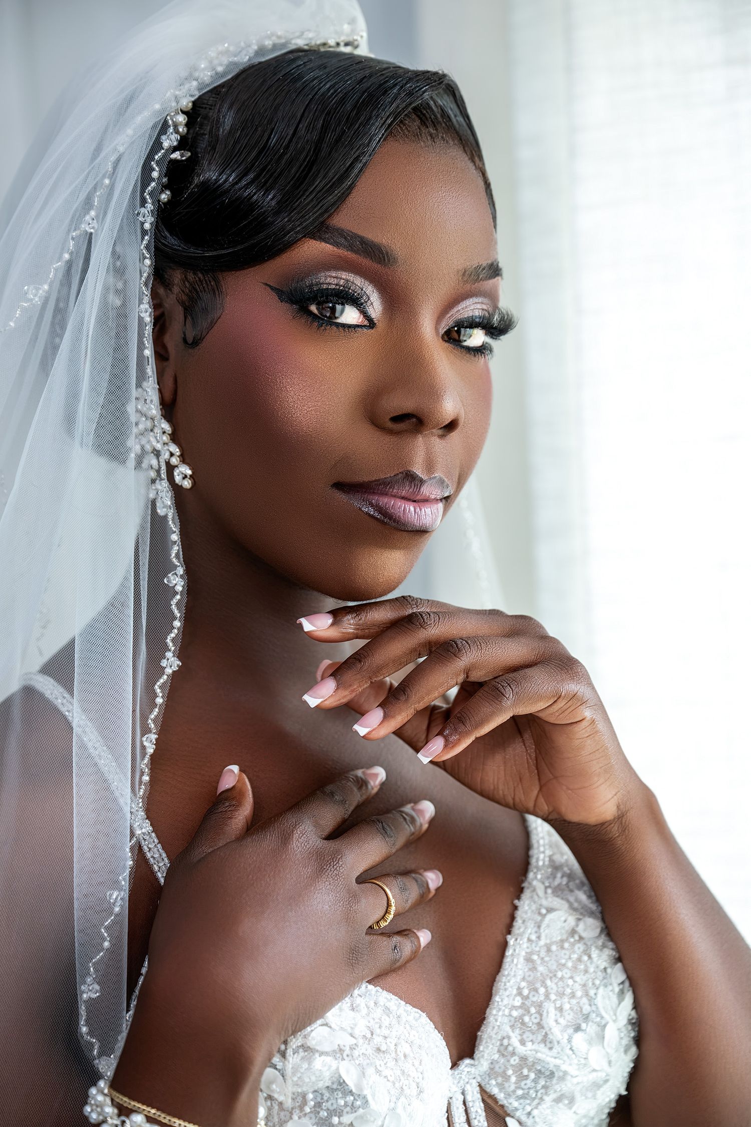 Elegant Black bride in white lace gown and veil, wearing rhinestone hair accessories and glam makeup, posing gracefully.