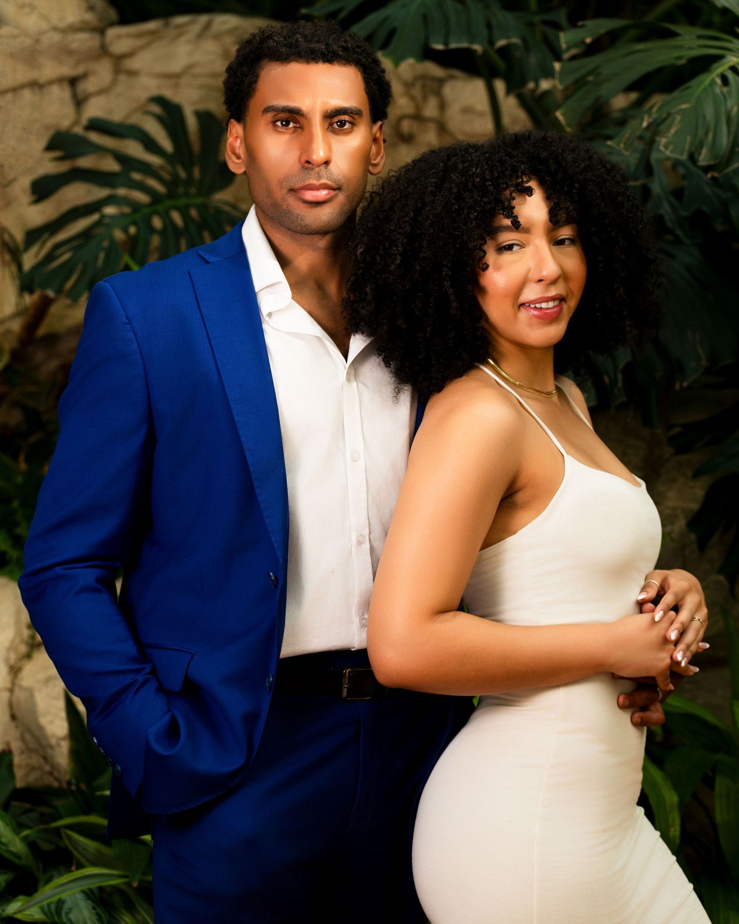 Elegant couple in formal attire pose together against tropical foliage backdrop.