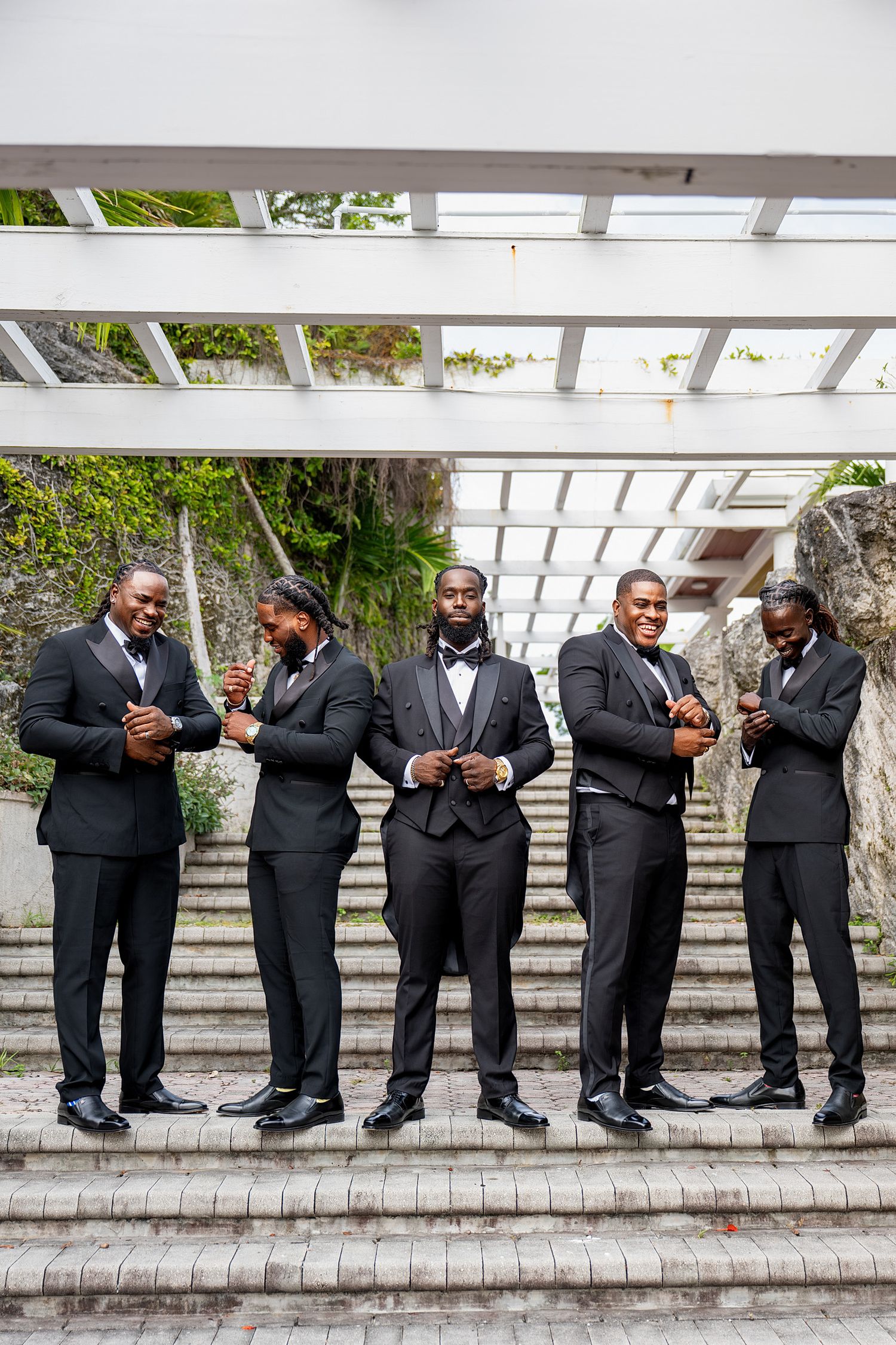 Five groomsmen in black tuxedos pose on stone steps under a white pergola at an outdoor wedding venue.