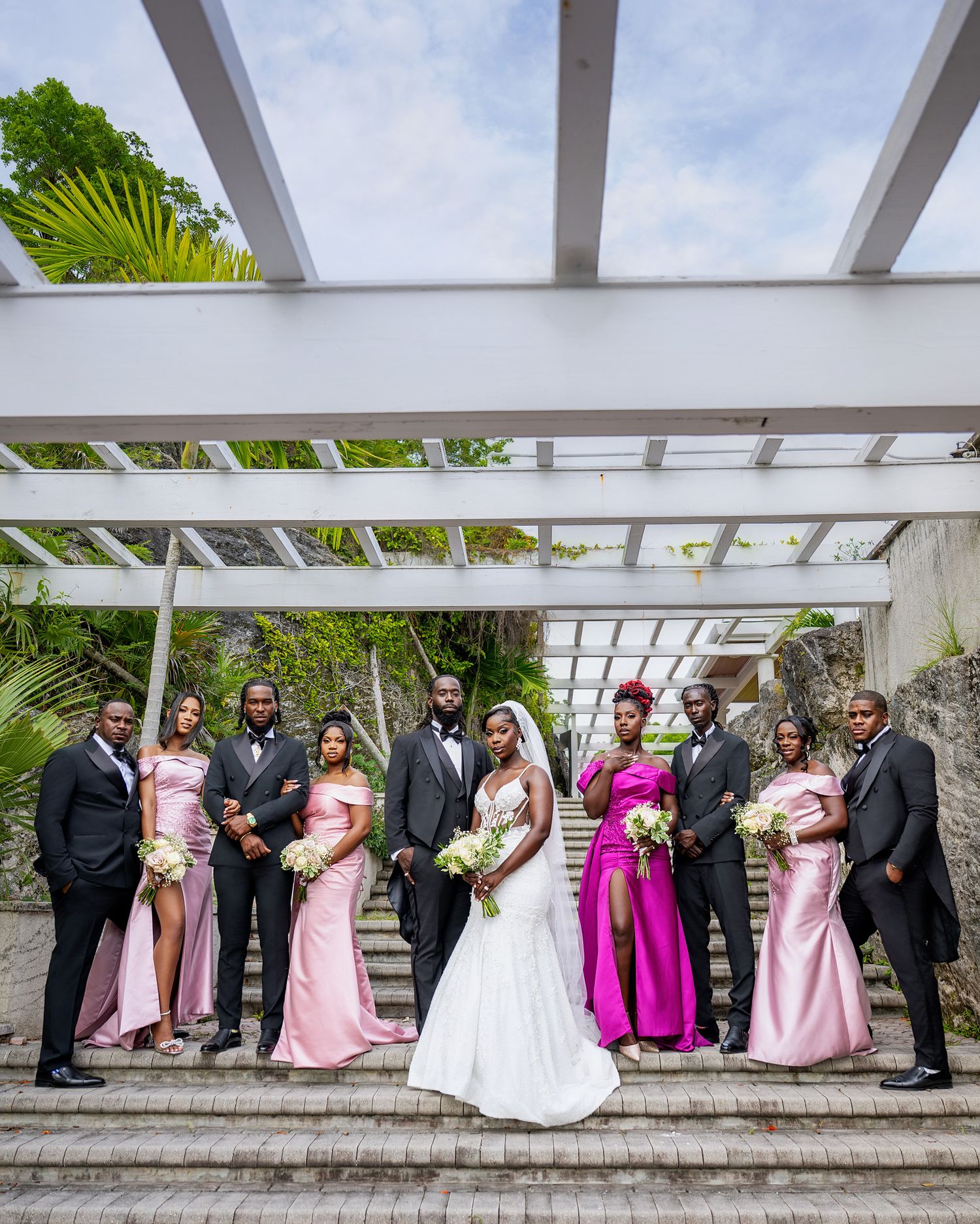 Wedding party poses on steps, bride in white gown, bridesmaids in pink and fuchsia dresses, groomsmen in black tuxedos.