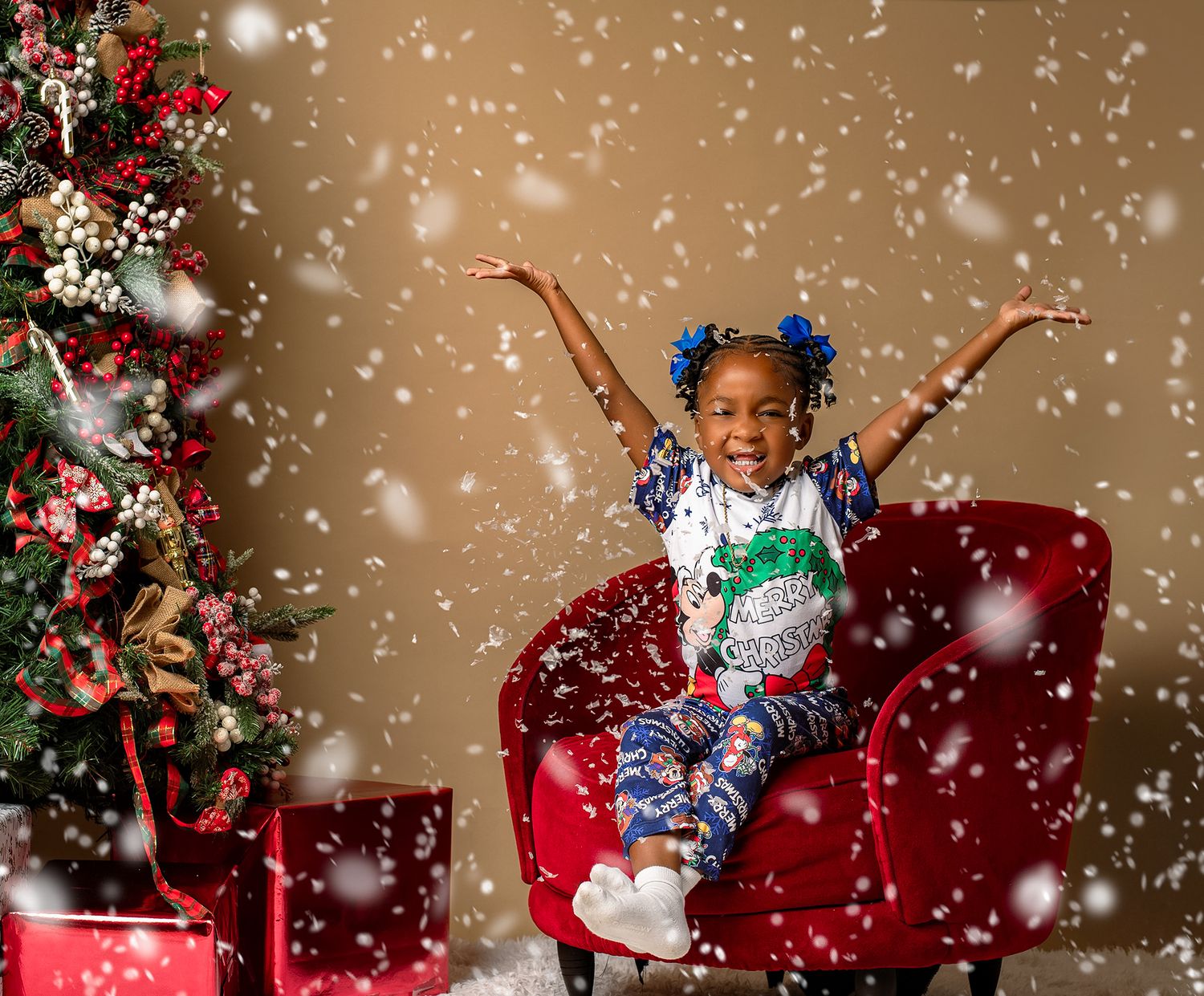 A joyful child jumps on a red chair surrounded by falling snow and Christmas decorations.