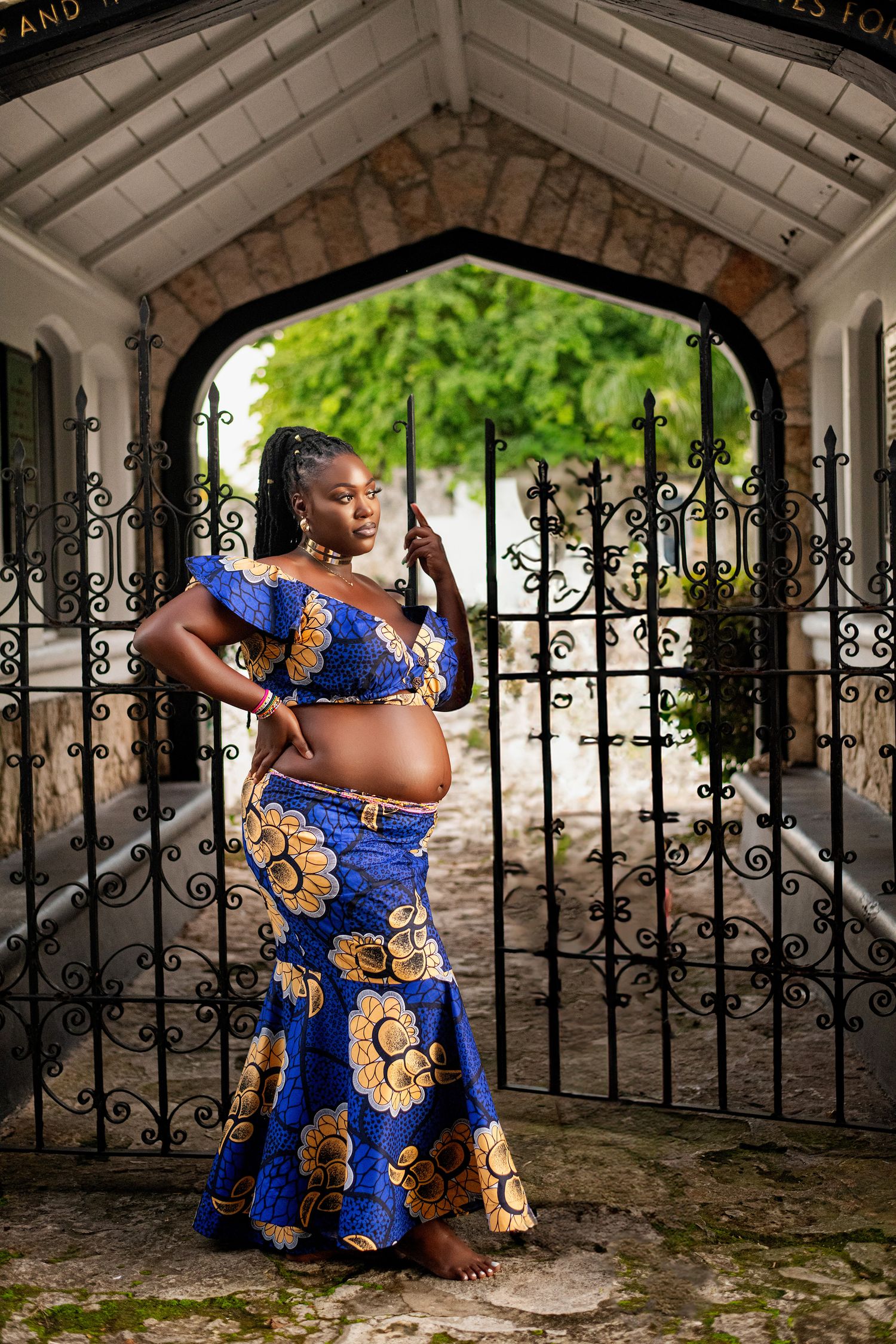 An expectant mother poses in vibrant blue and gold traditional African attire by an ornate iron gate.