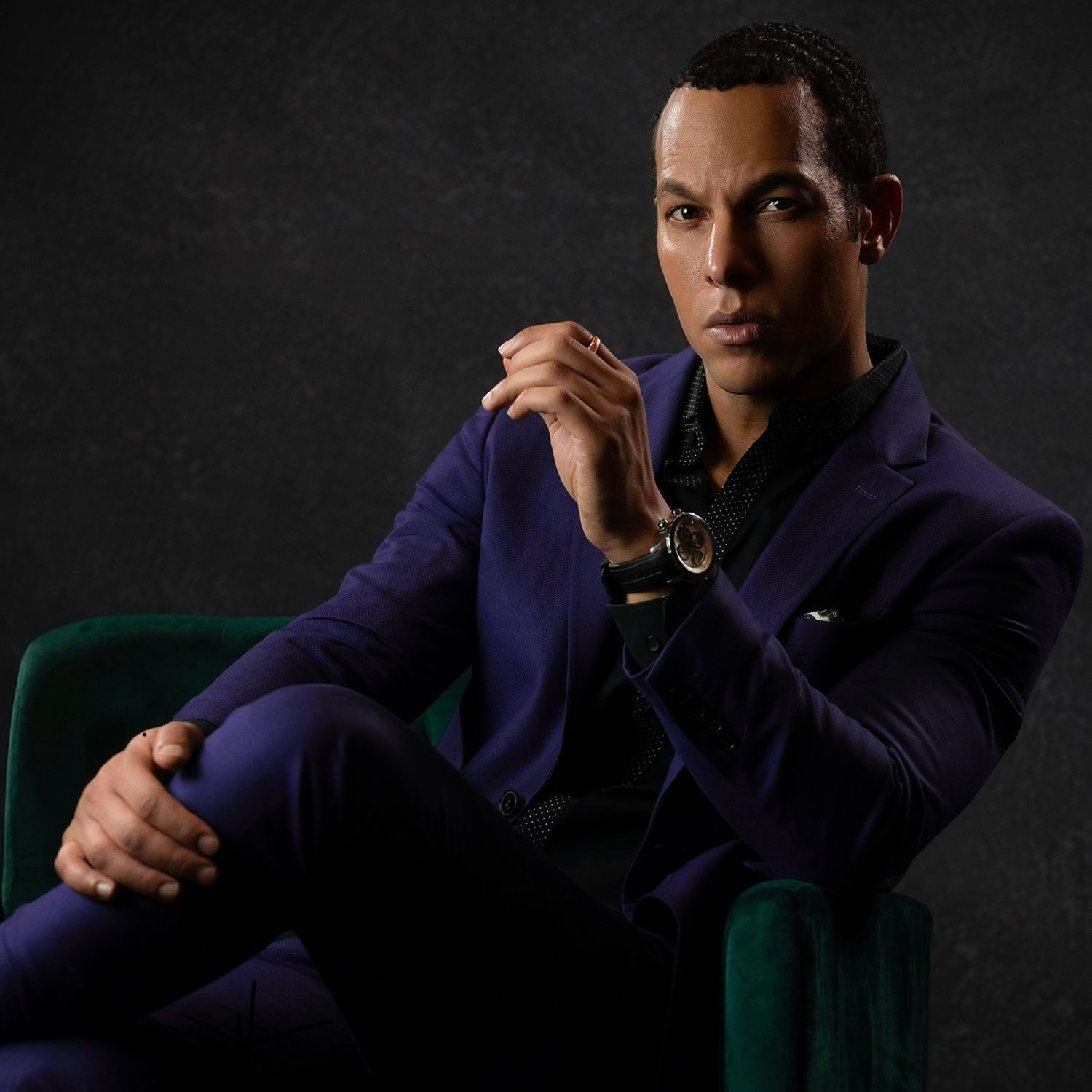 Professional studio portrait of man in blue suit jacket seated in green armchair.