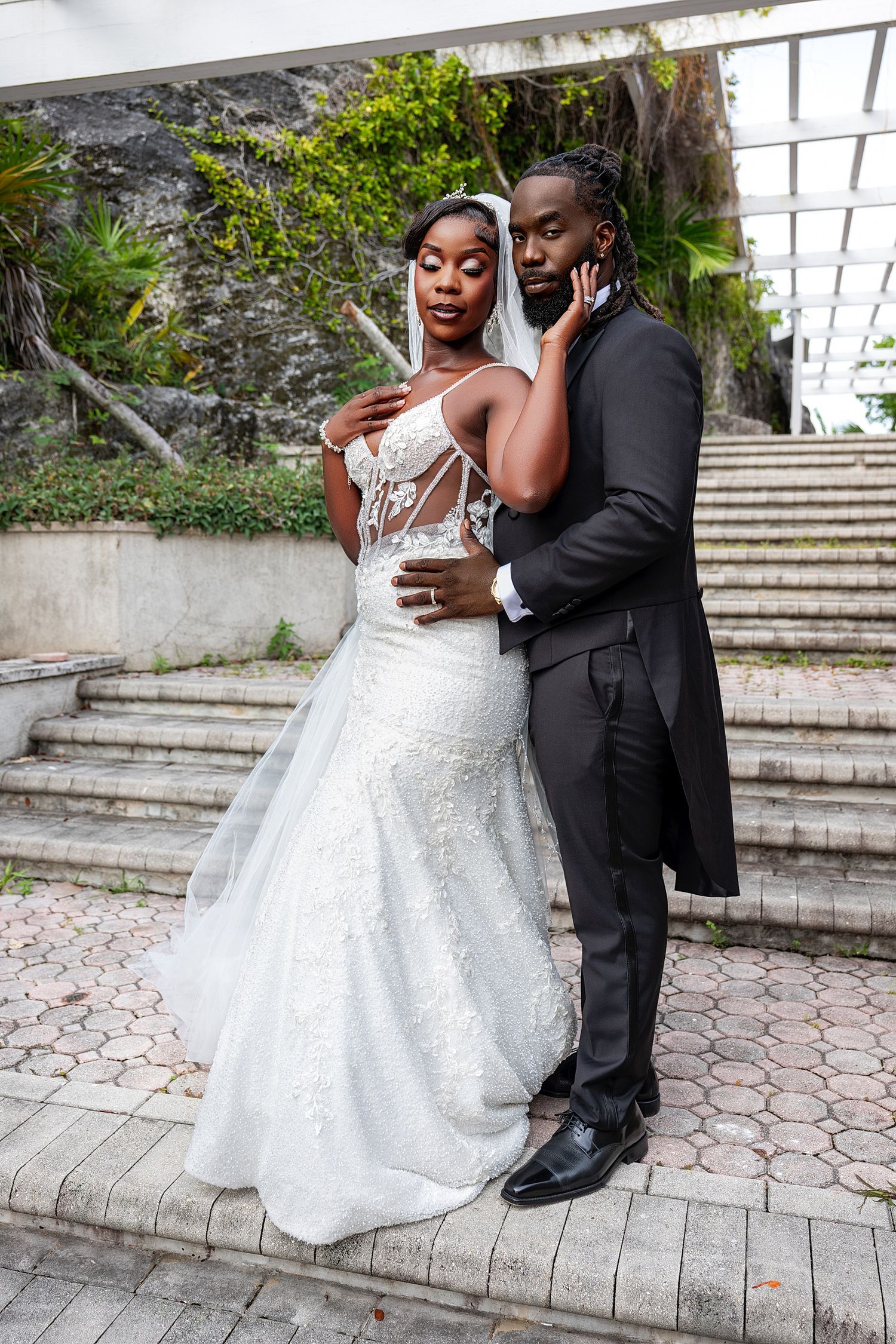 Elegant Black couple in wedding attire pose on stone steps, bride in white lace gown with veil, groom in black tuxedo.