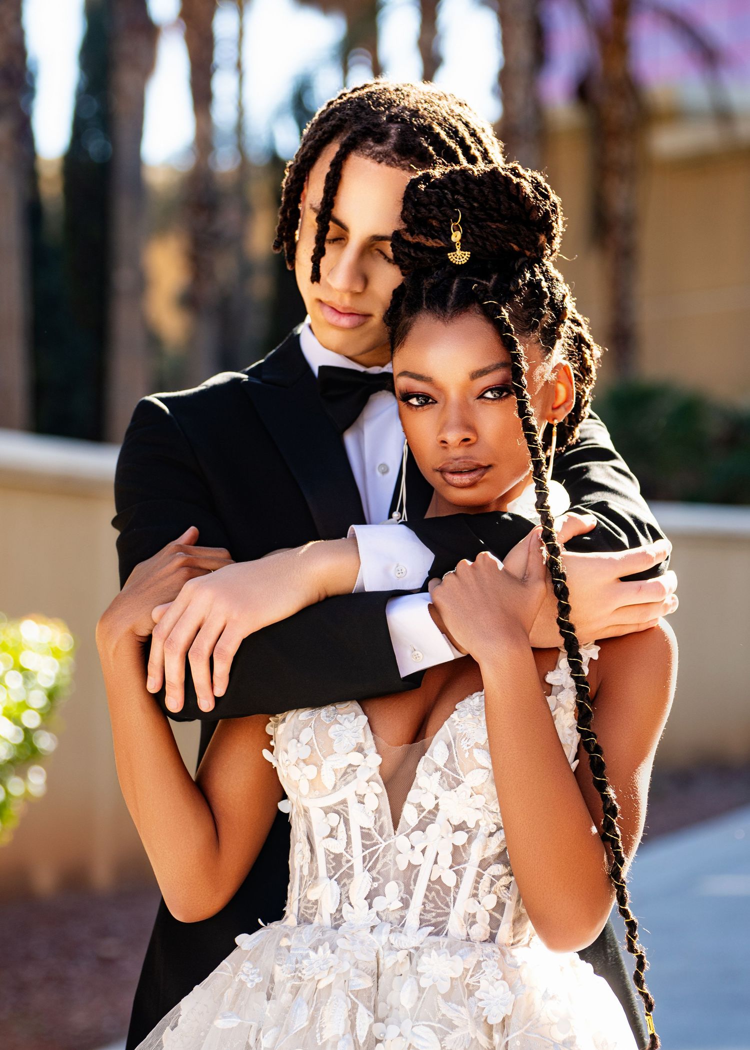 Formal couple embrace in black tuxedo and white lace wedding dress outdoors.