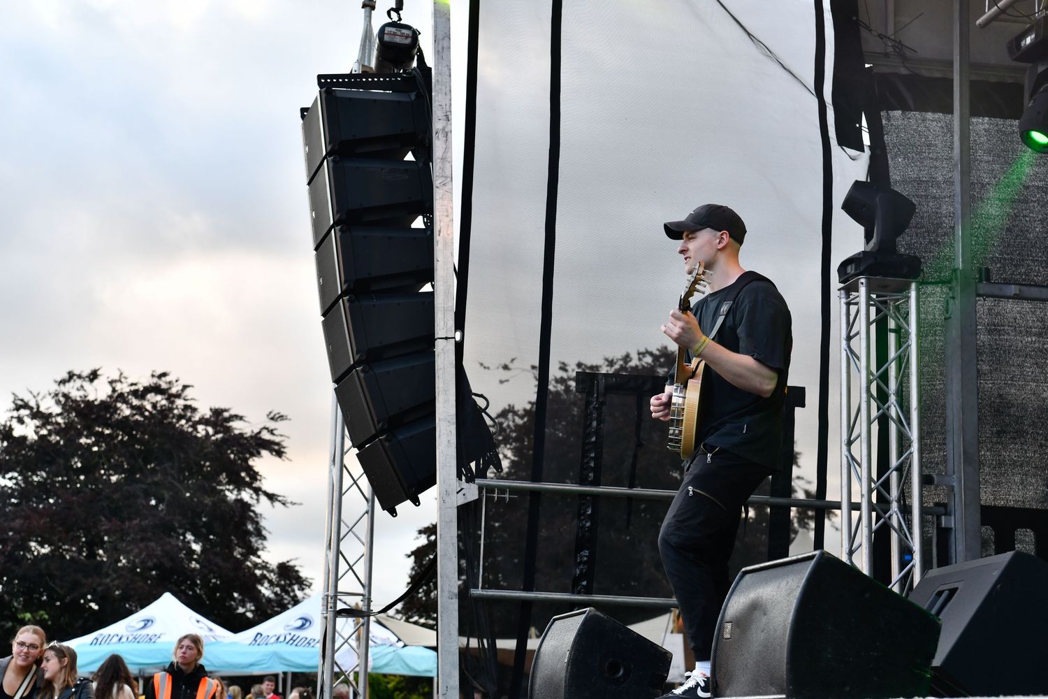Performers play on a large outdoor concert stage with lighting equipment and tents visible in the background.
