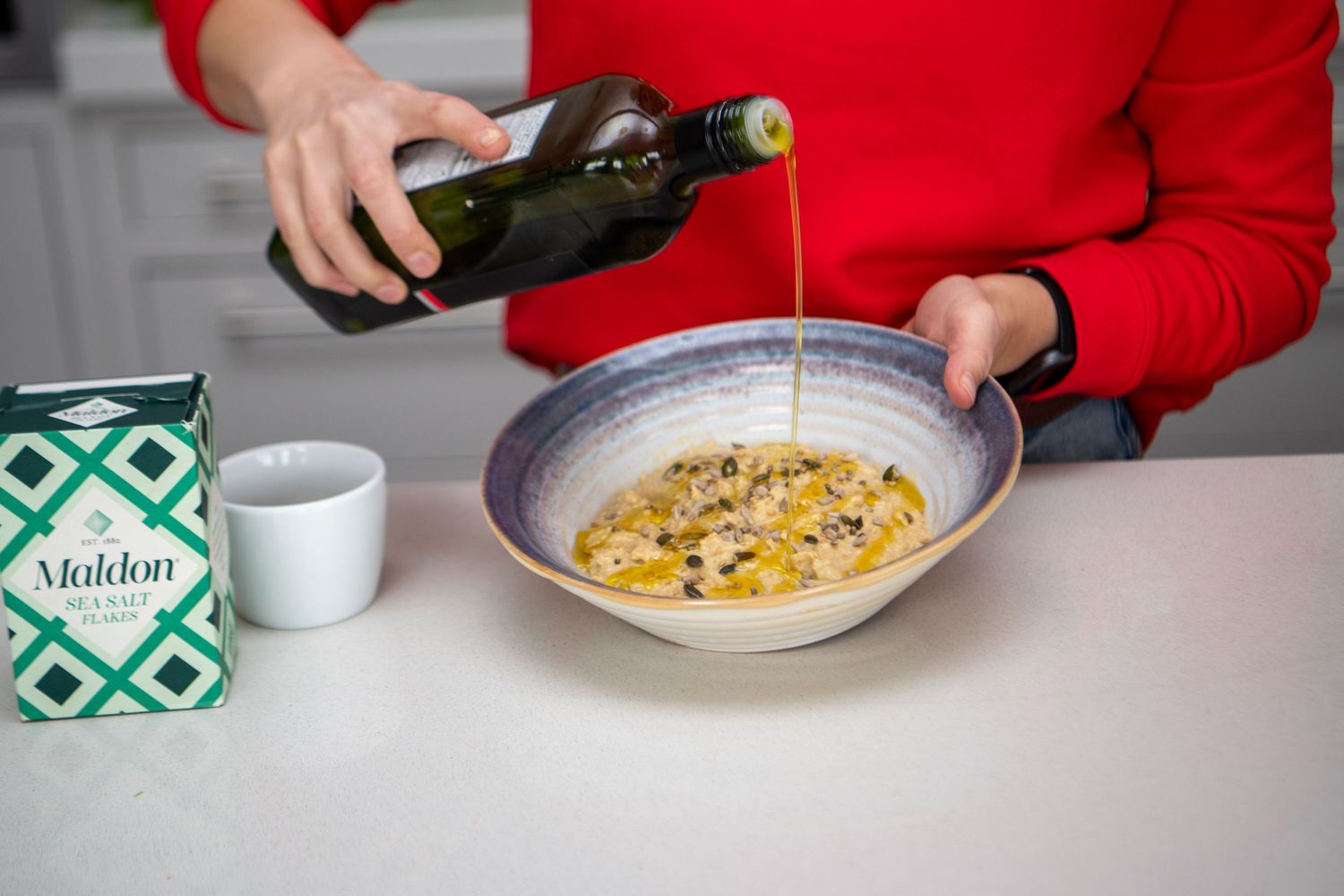 Person in red sweater pouring olive oil over a bowl of creamy soup.