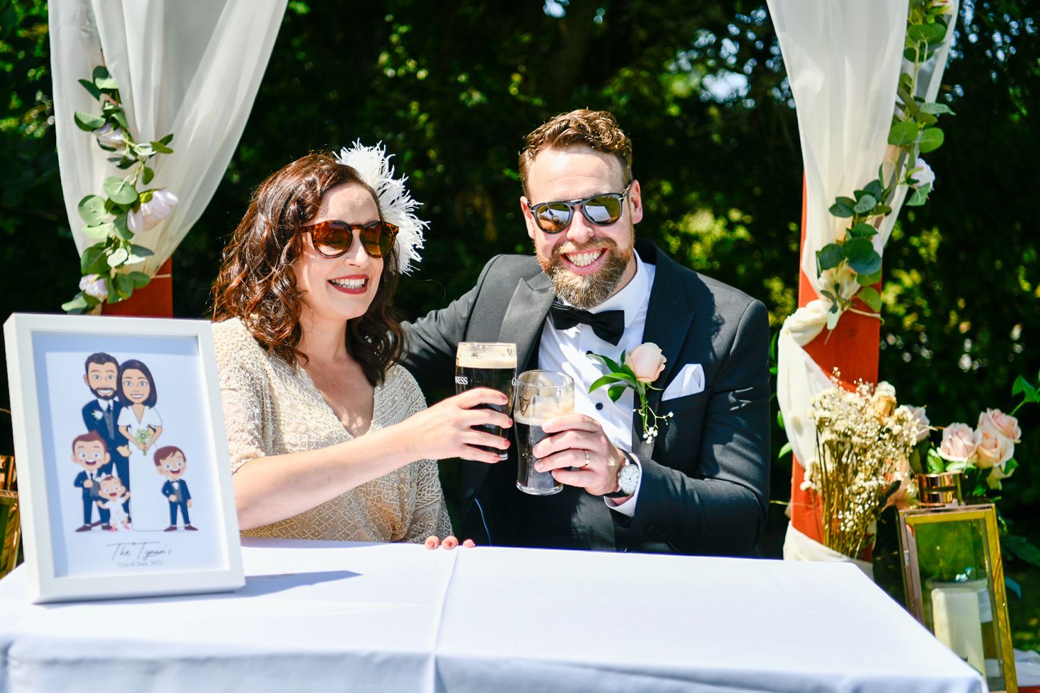 A couple toasting drinks at an outdoor wedding ceremony table with white drapes and a framed photo display.