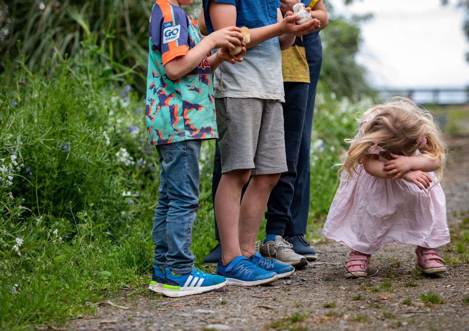 Children stand on a nature path while one crouches down among the tall grass.