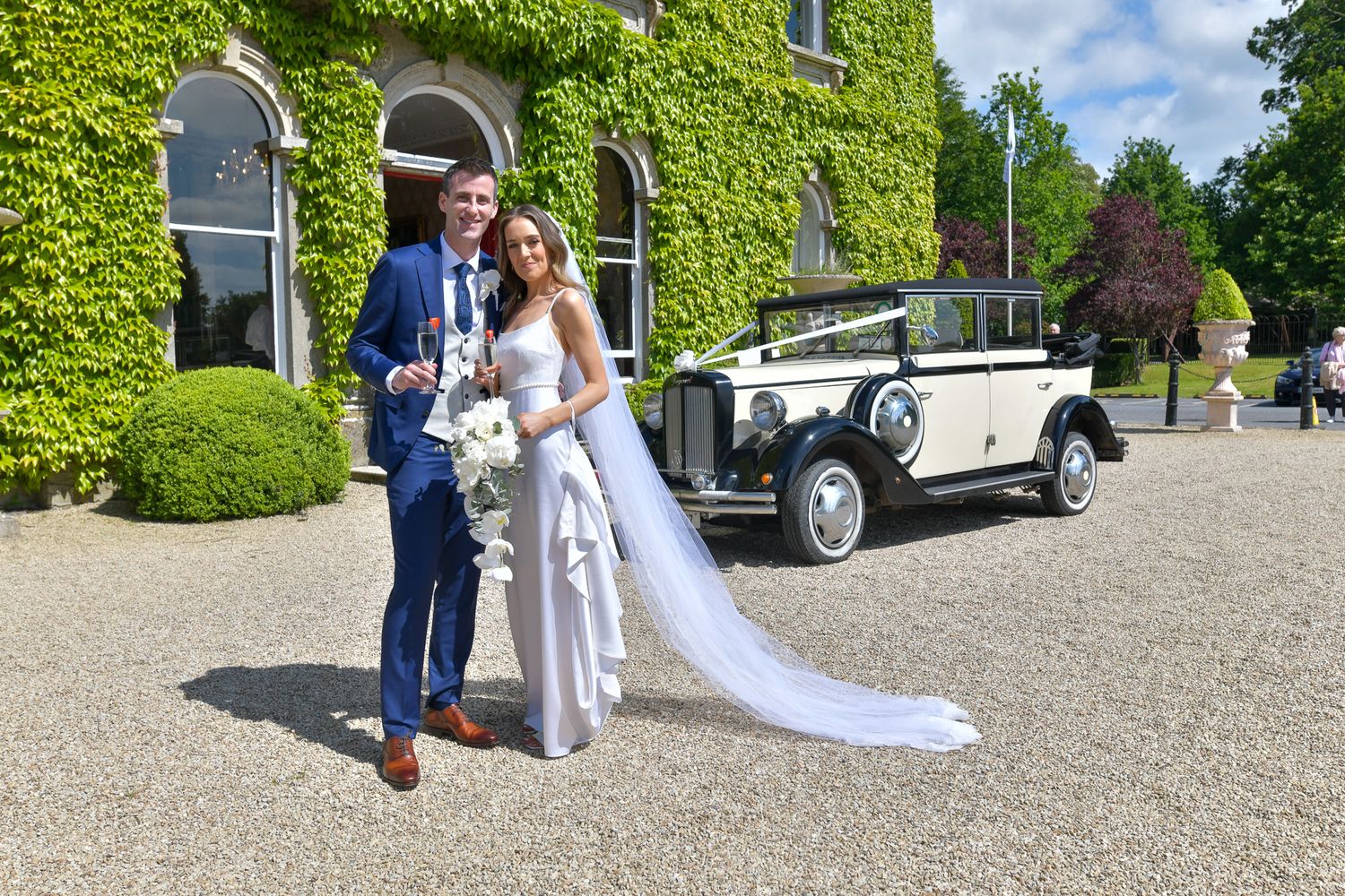 A couple poses in front of an ivy-covered building with a vintage car in the background.