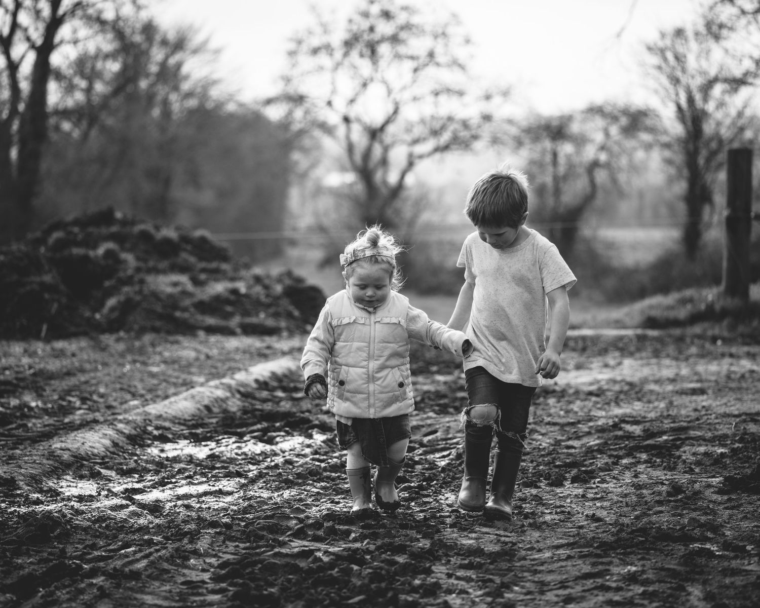 Two small figures walk hand in hand through a misty winter landscape along a muddy rural path.
