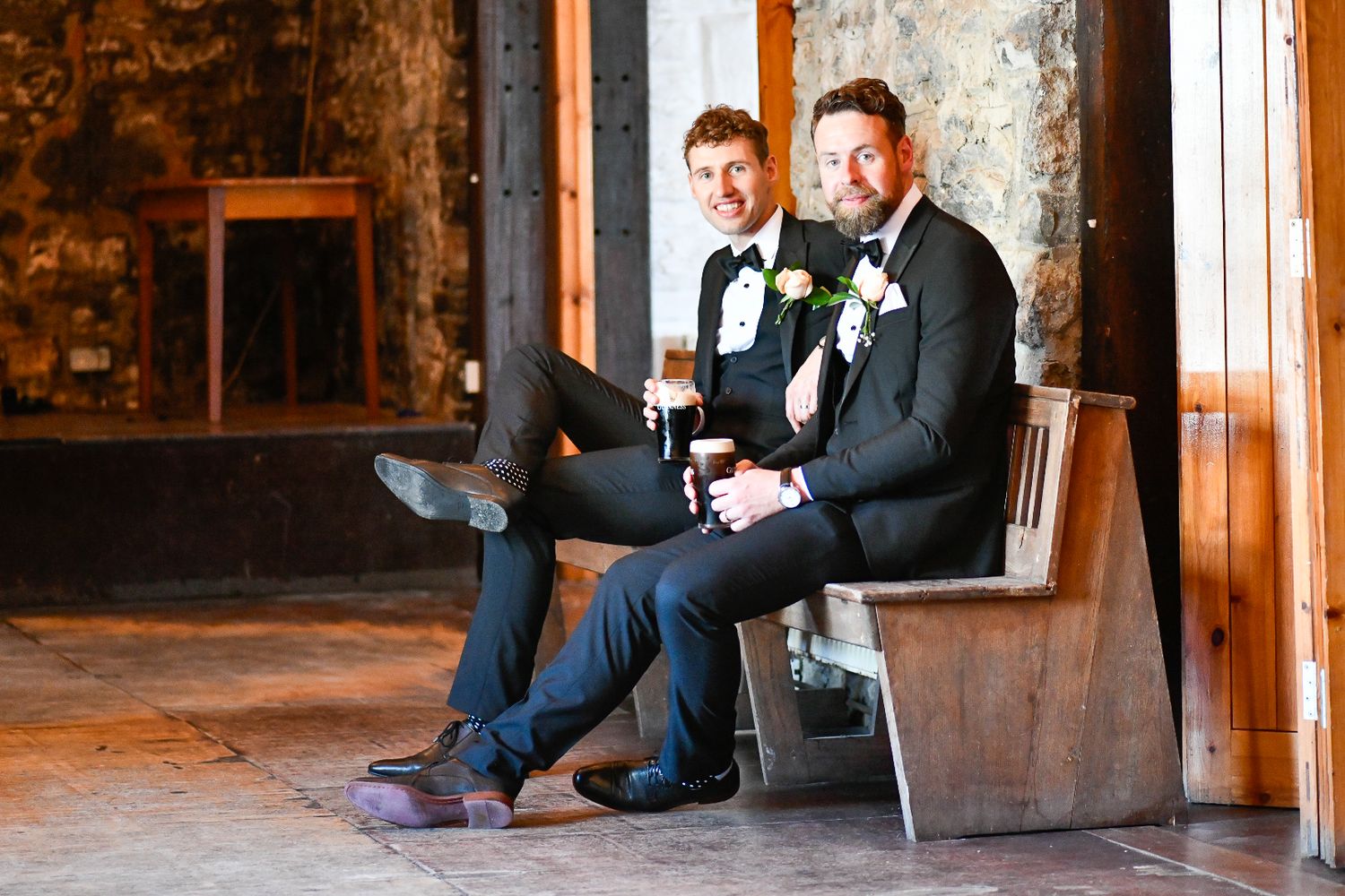 Two people in formal attire sitting on bench drinking beverages in rustic indoor setting.