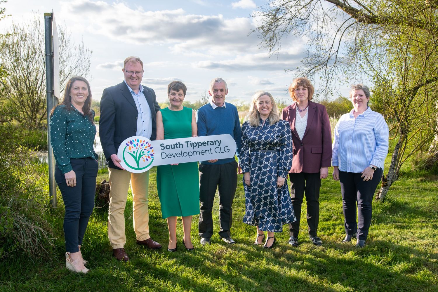 A group poses outdoors with a green university sign on a sunny spring day.