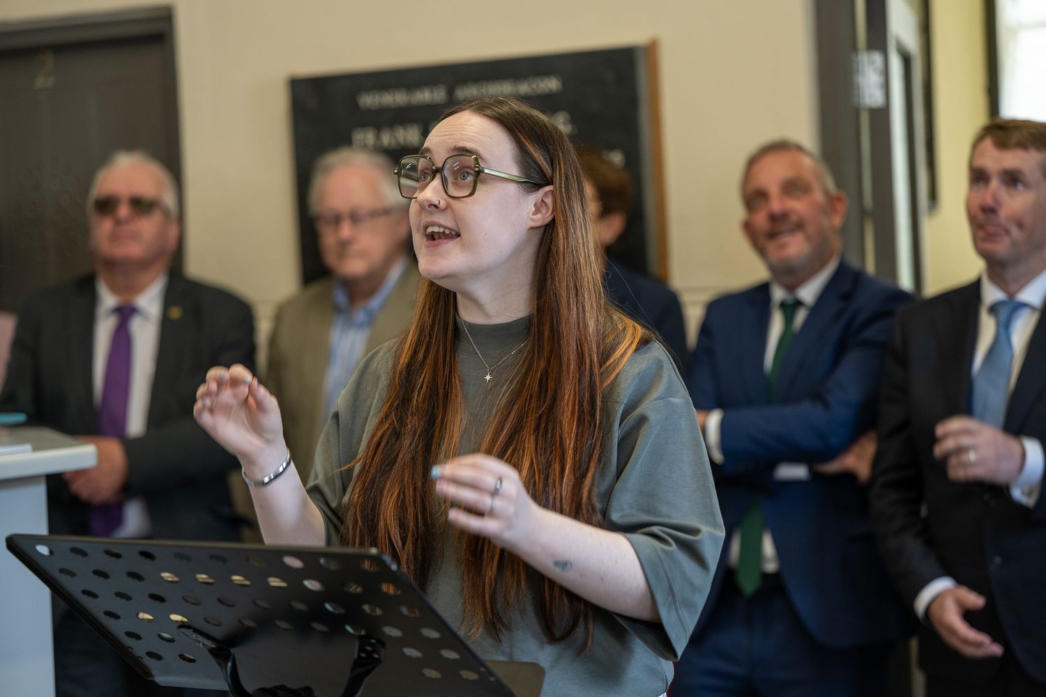 A speaker with long red hair gives a presentation while several business people watch from behind.