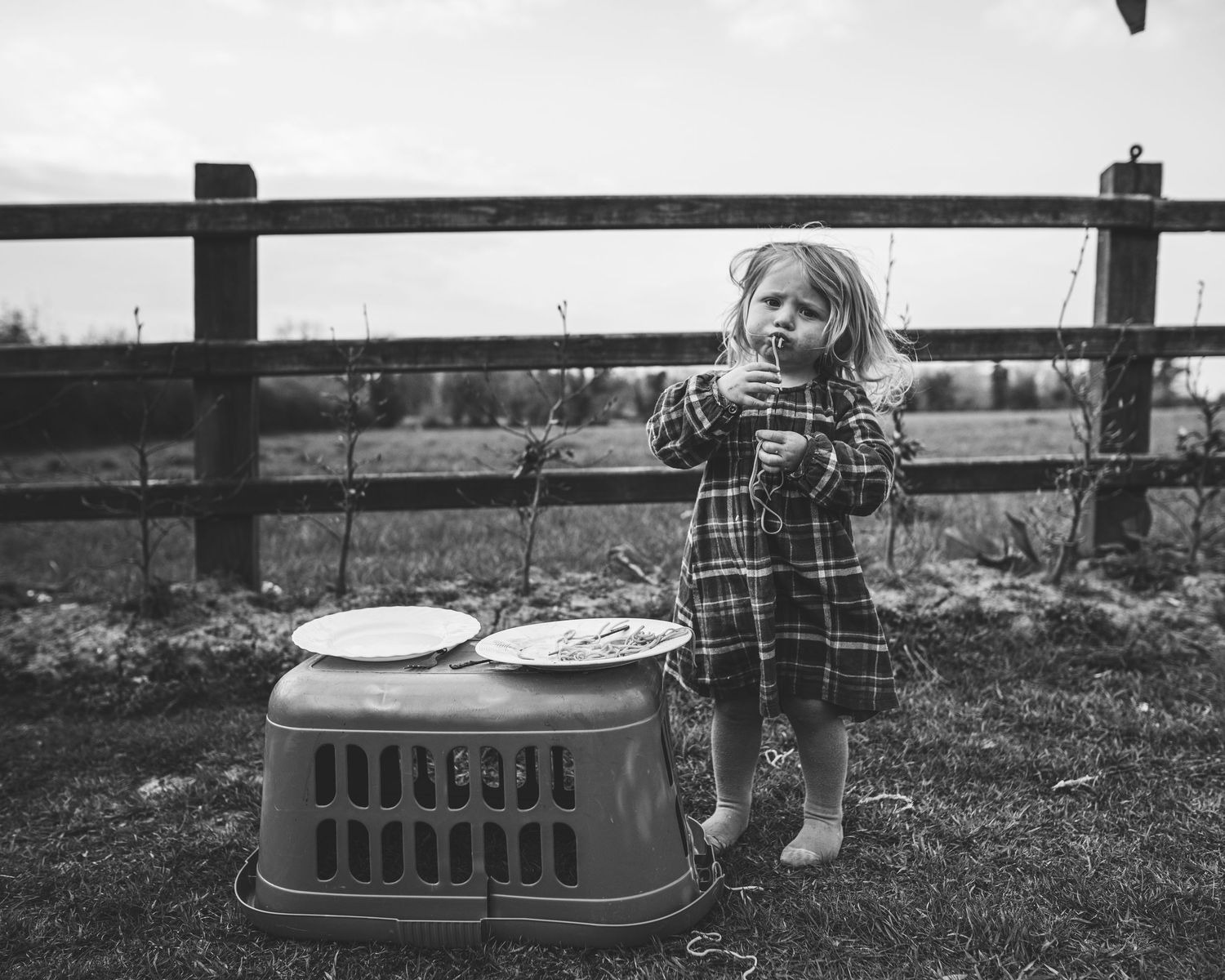 Vintage black and white photo showing a young figure standing next to a pet carrier outdoors.