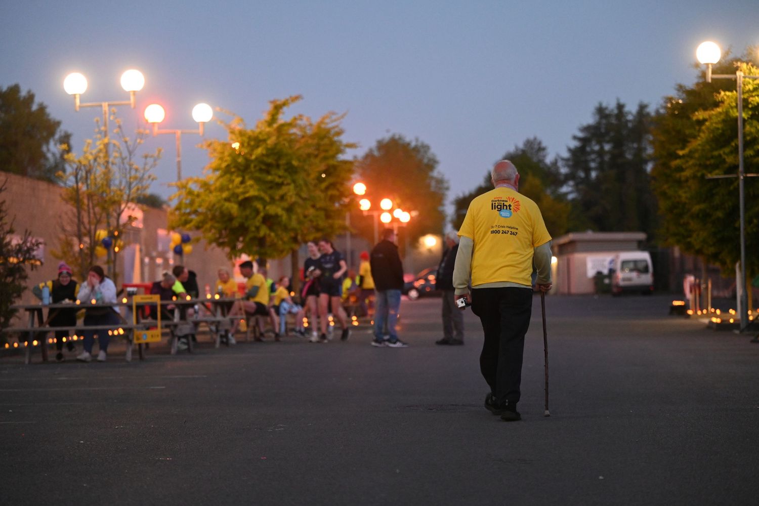A person in yellow walks with a cane at dusk as street lights glow in the background.