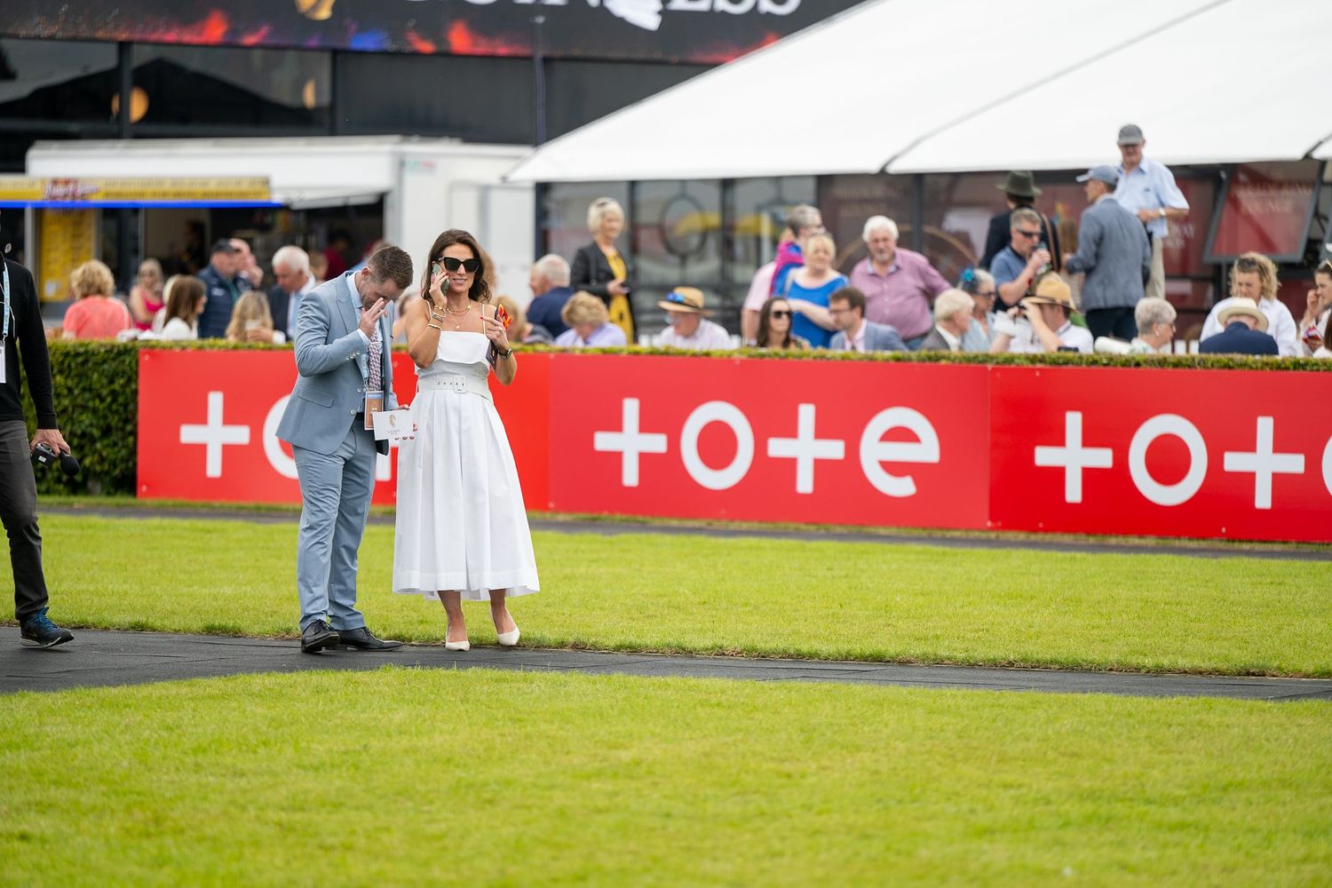 Outdoor event with crowd gathering near advertising banners on grassy field.