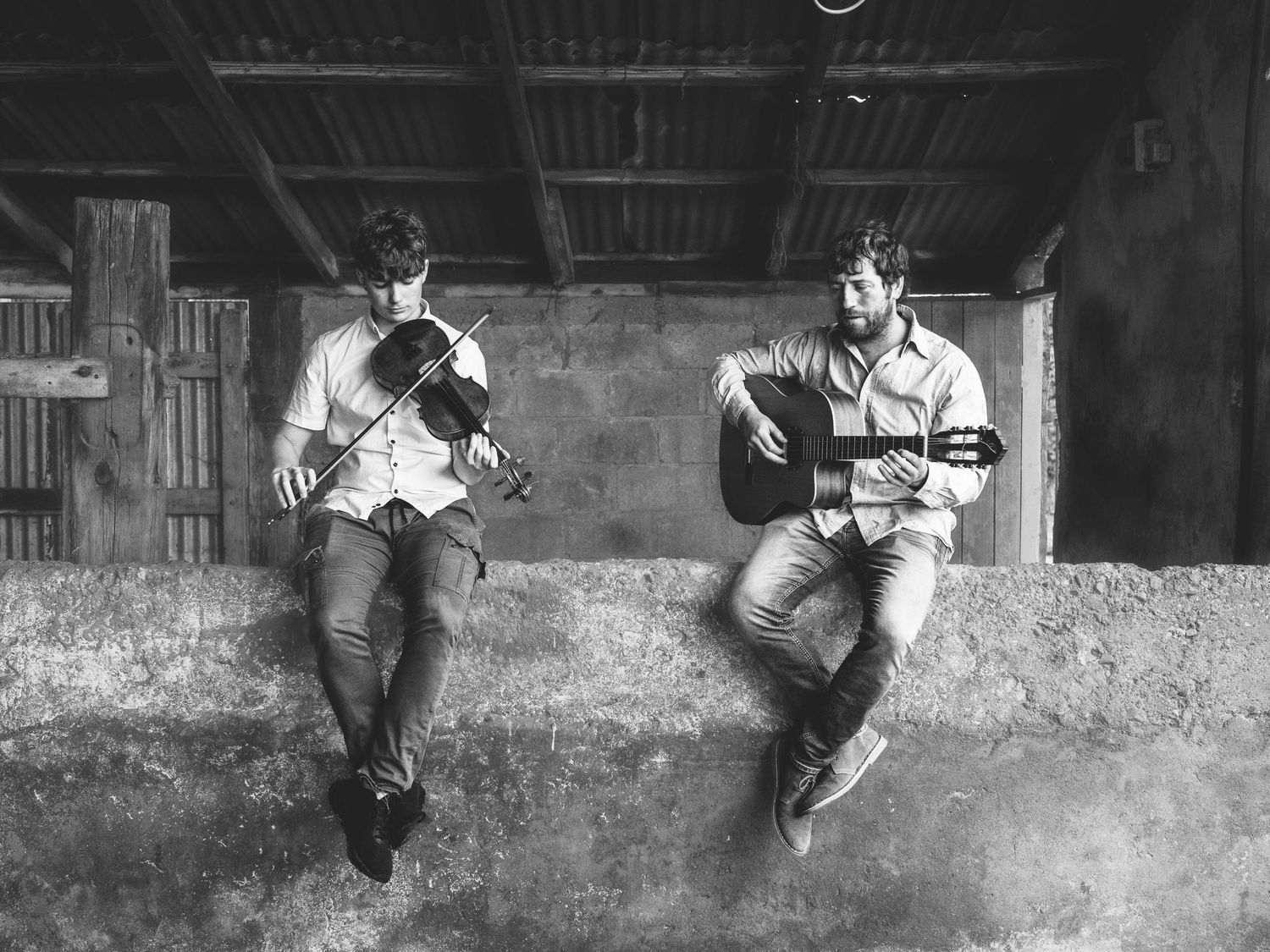 Two musicians sit casually on a ledge playing acoustic guitars in a rustic black and white setting.