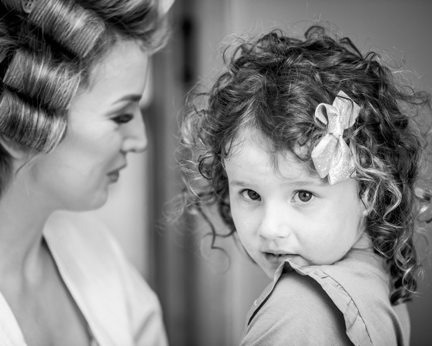 Black and white portrait of curly-haired toddler wearing hair flower with someone in hair rollers.