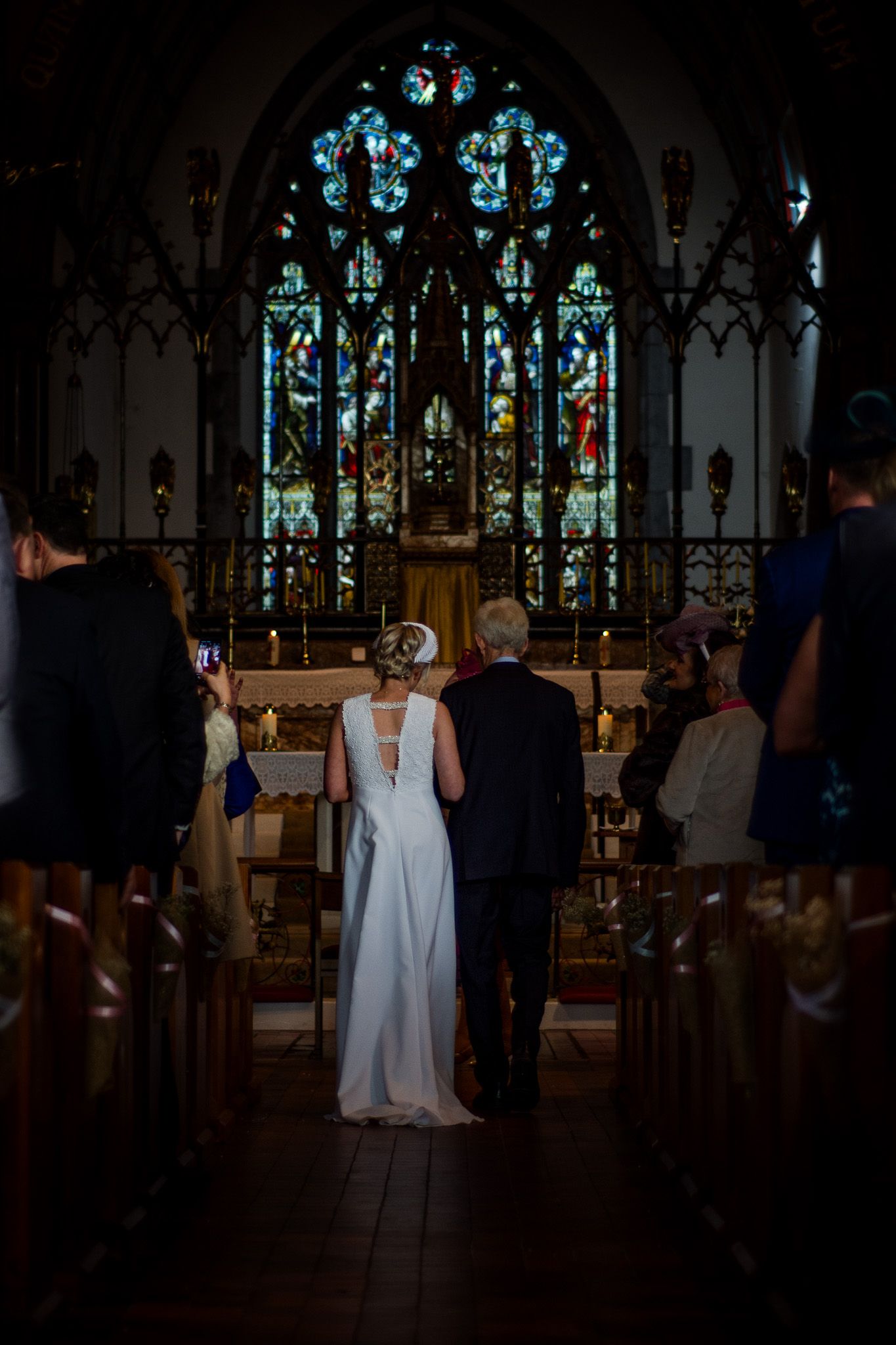 A bride in a white dress stands before a stunning stained glass church window during a ceremony.