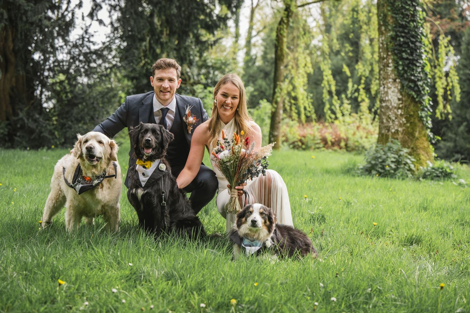 A couple sits on grass with their three dogs in a lush garden setting.