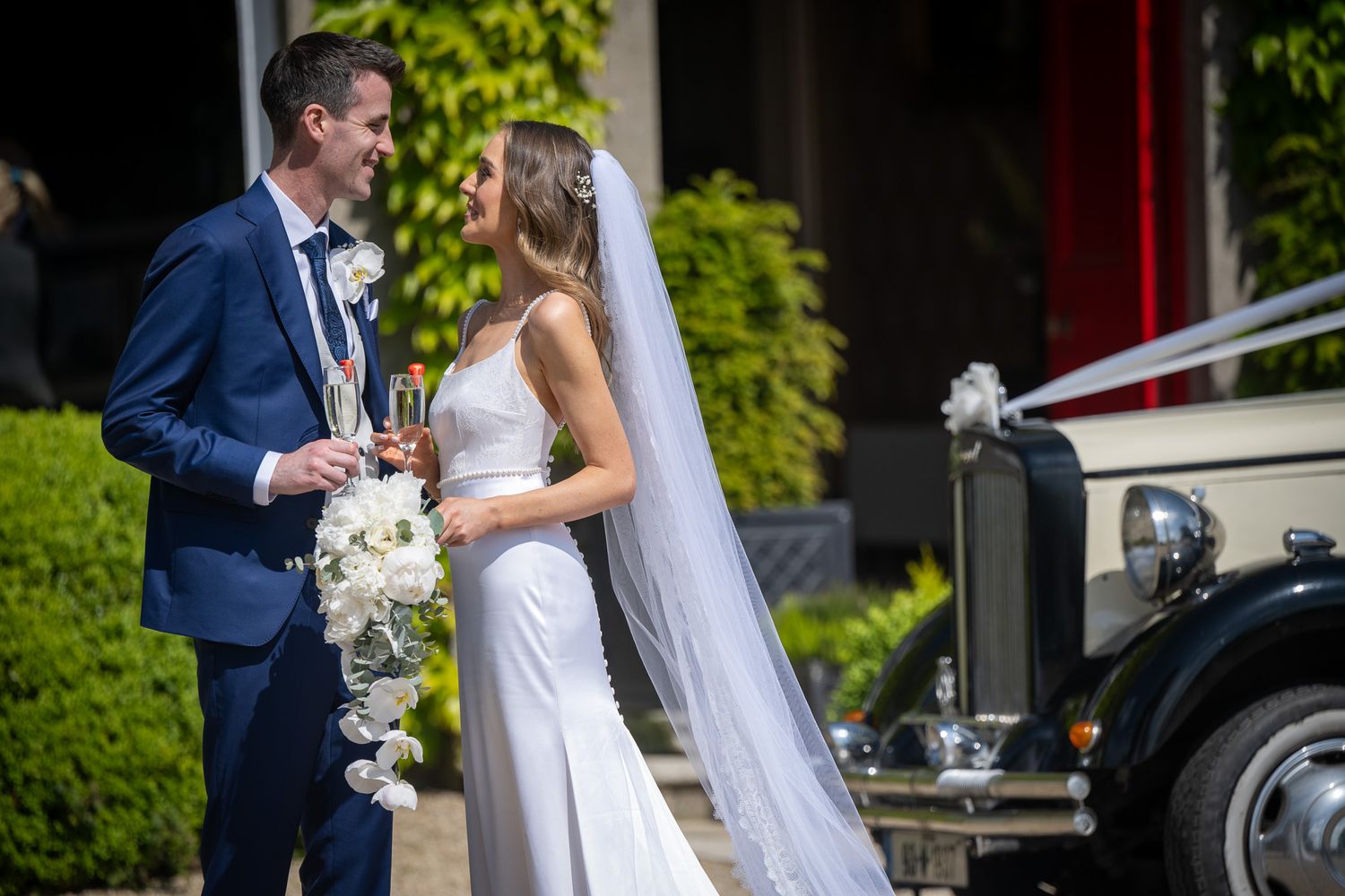A couple shares champagne outdoors near a classic car at their wedding venue.