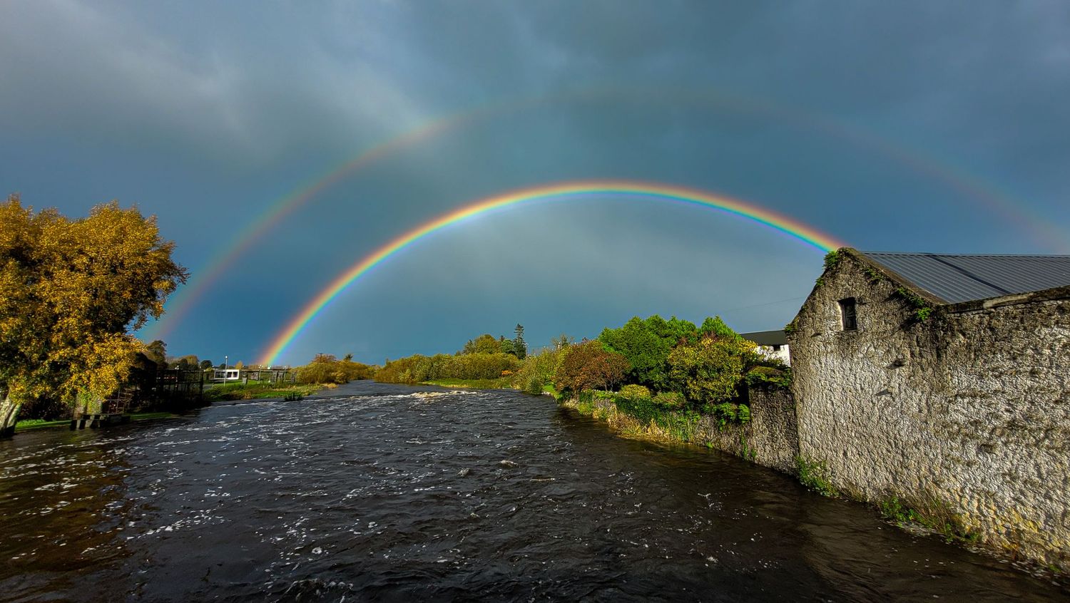Double rainbow arches over a river and stone building under dramatic stormy skies.