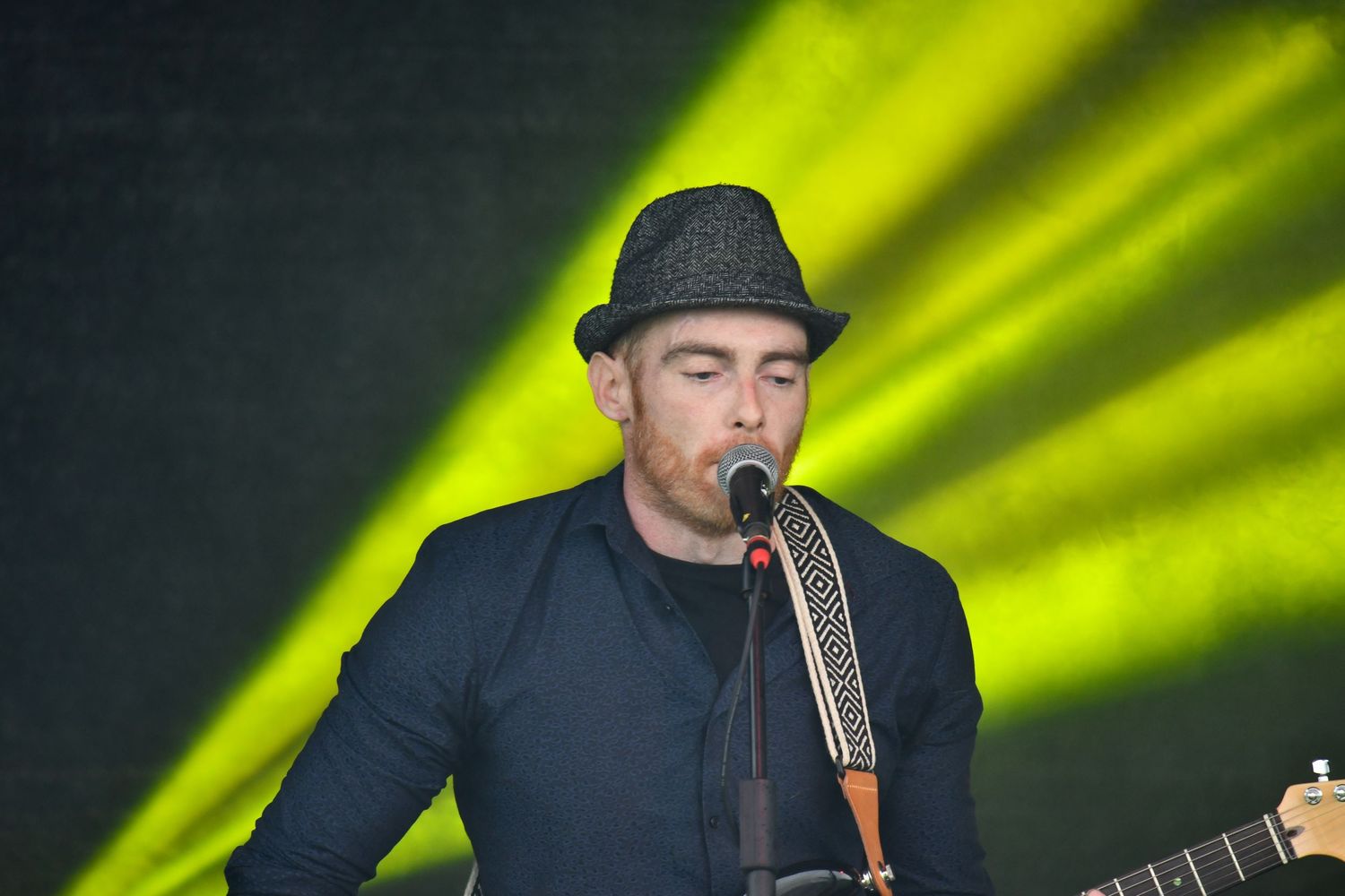 A musician in a dark outfit and fedora performs with a guitar against green stage lighting.