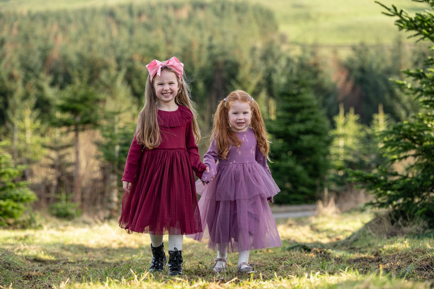 Two children in burgundy and purple dresses standing among evergreen trees.