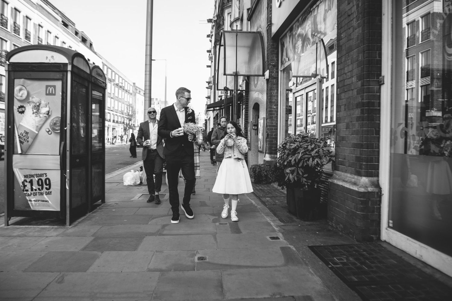 Black and white street scene with people walking past shops and a phone booth in an urban setting.