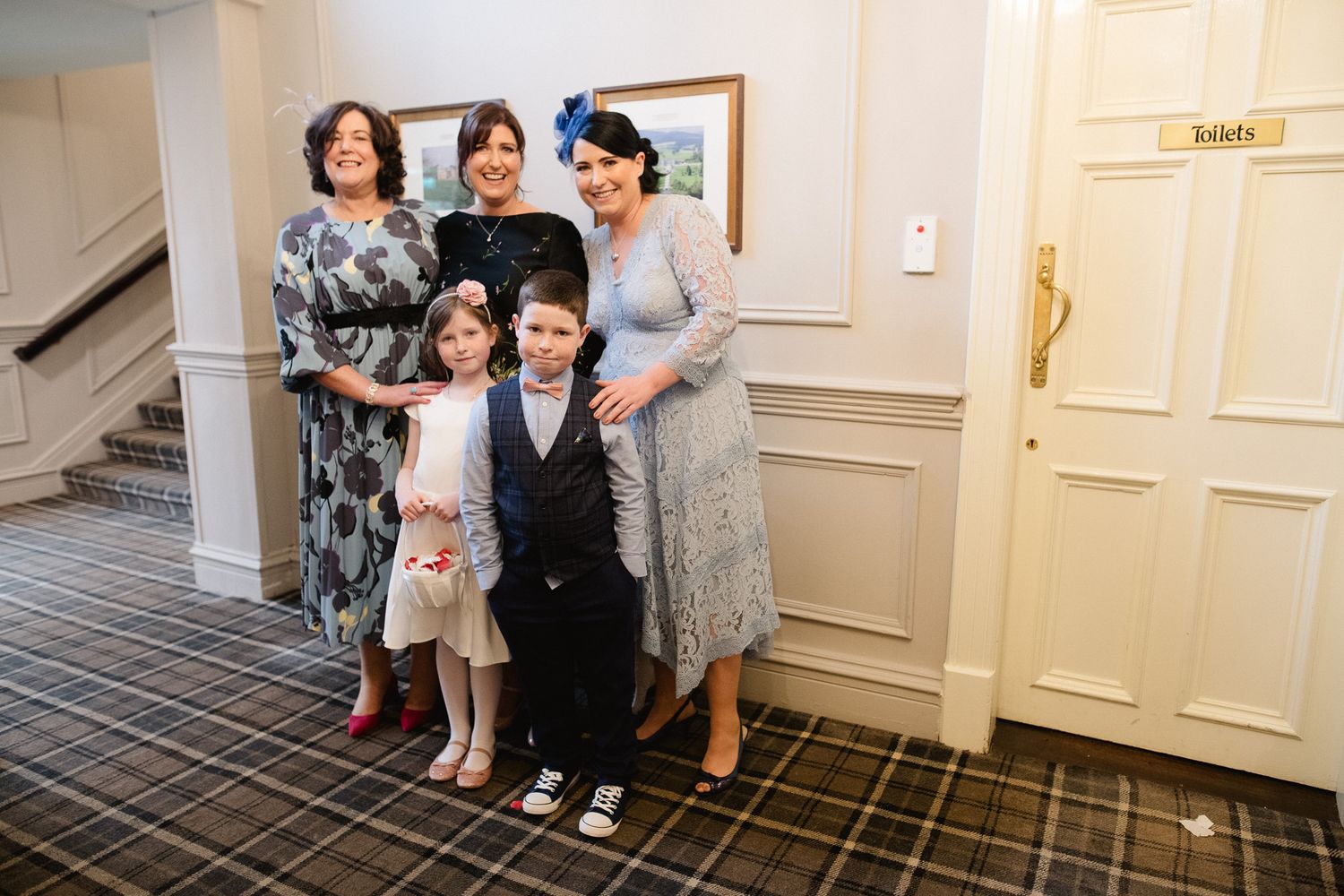 Wedding party members gather for a group photo in a hallway with patterned carpet.