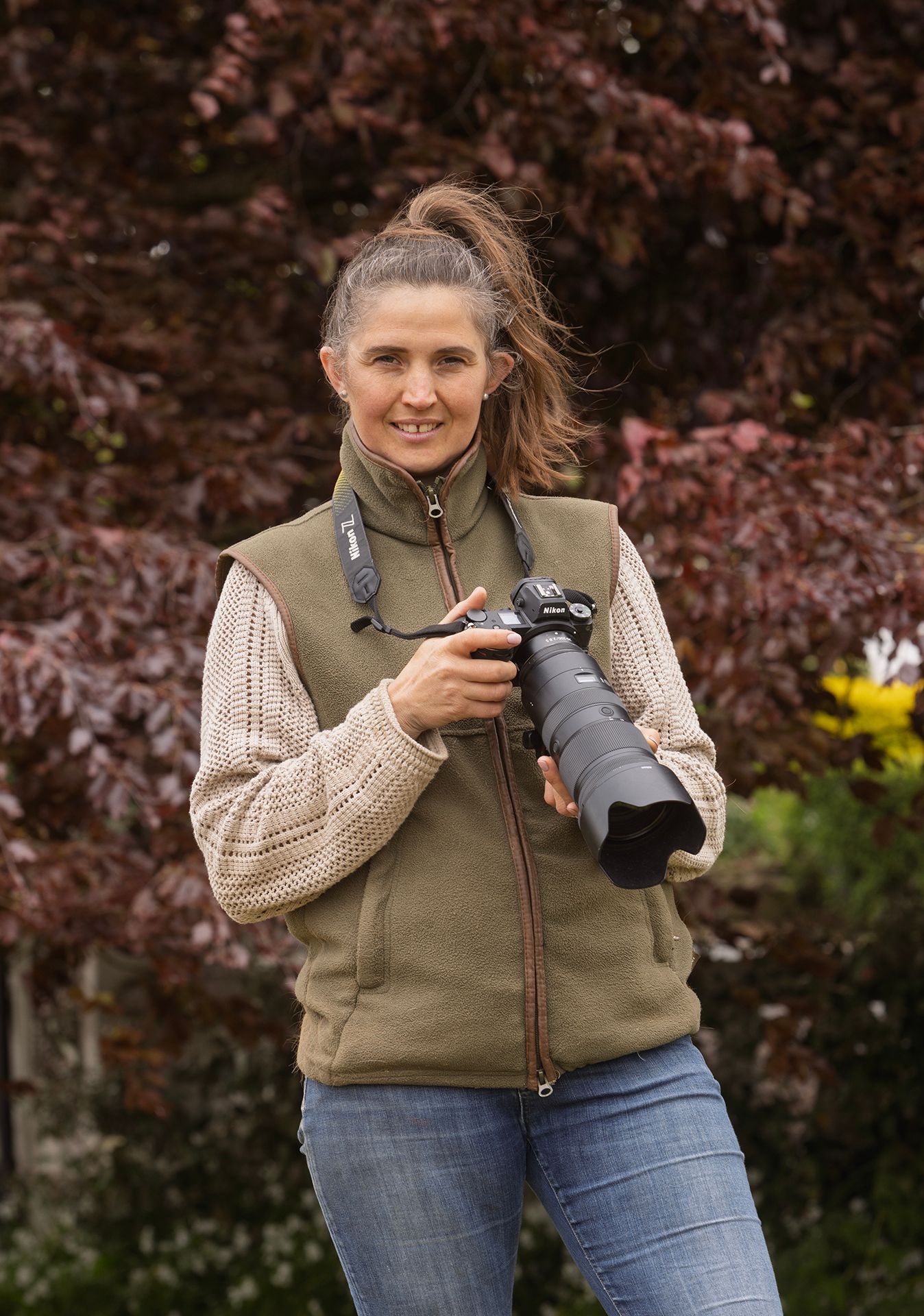 Nature photographer wearing a vest and sweater holds professional camera gear outdoors.