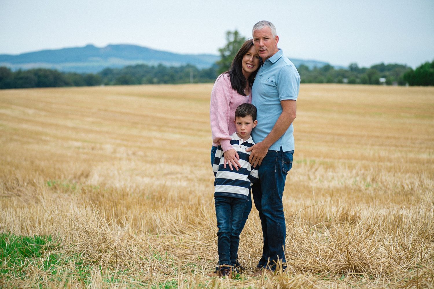 Three people embrace while standing in a golden wheat field with mountains in the background.