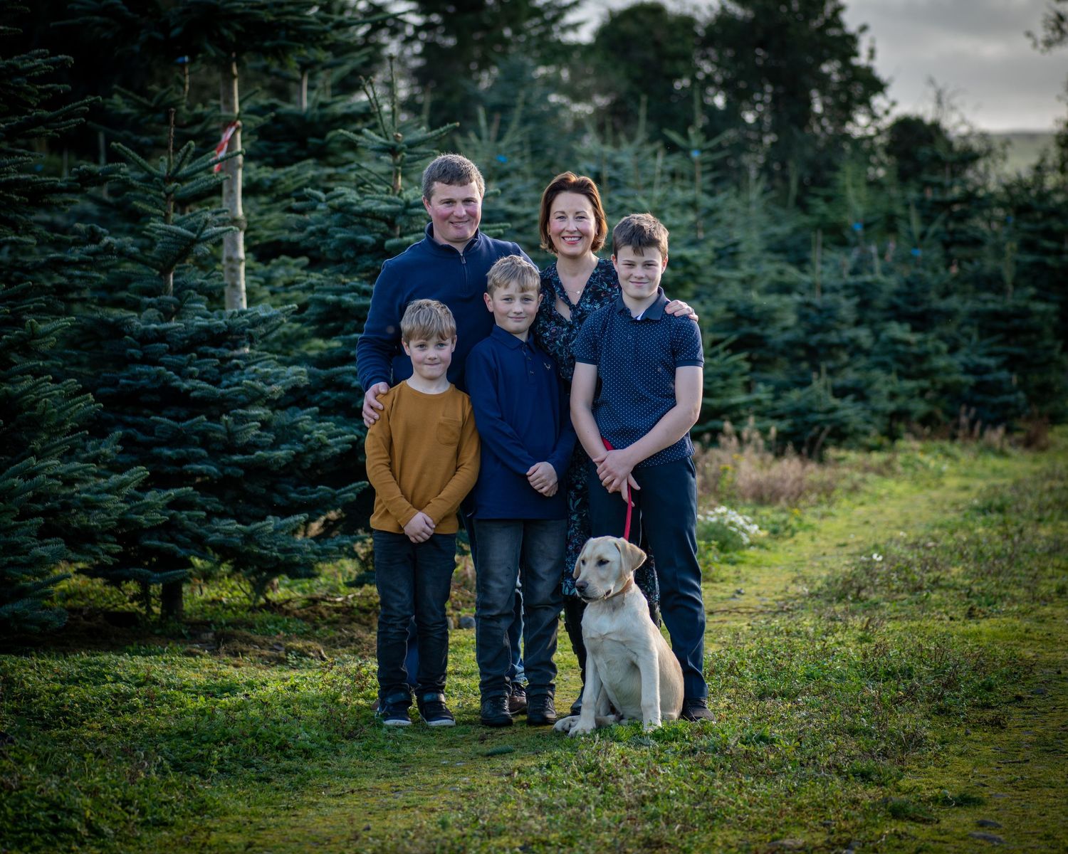 Family and pet dog posing together in a Christmas tree farm with evergreen trees in the background.