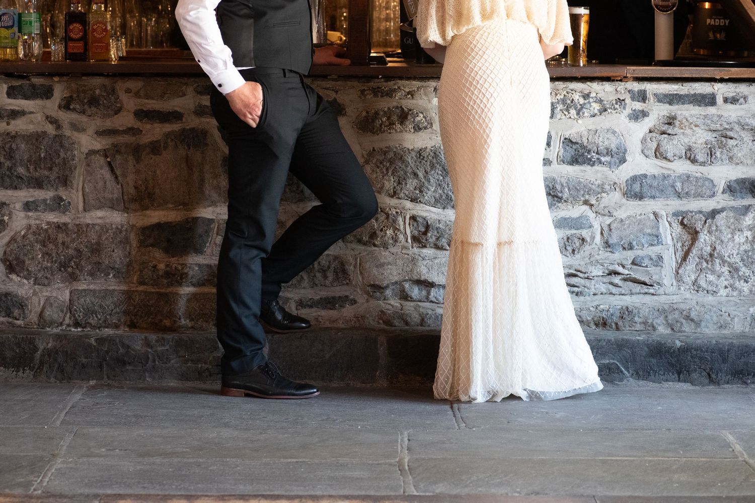 A formal couple's legs shown against a stone wall, with the bride wearing a flowing white dress.