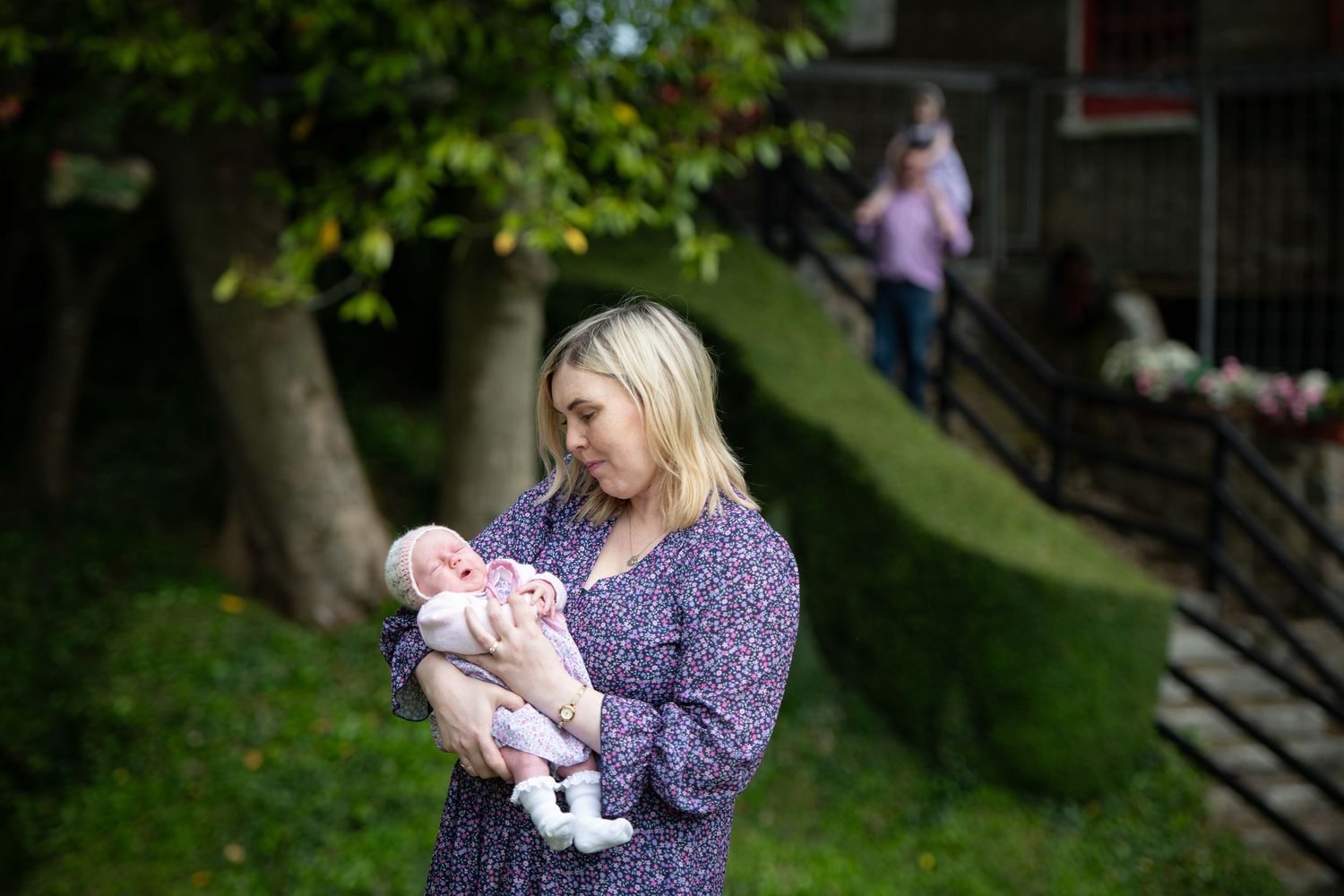 Woman in purple dress holding infant outdoors with lush green background.