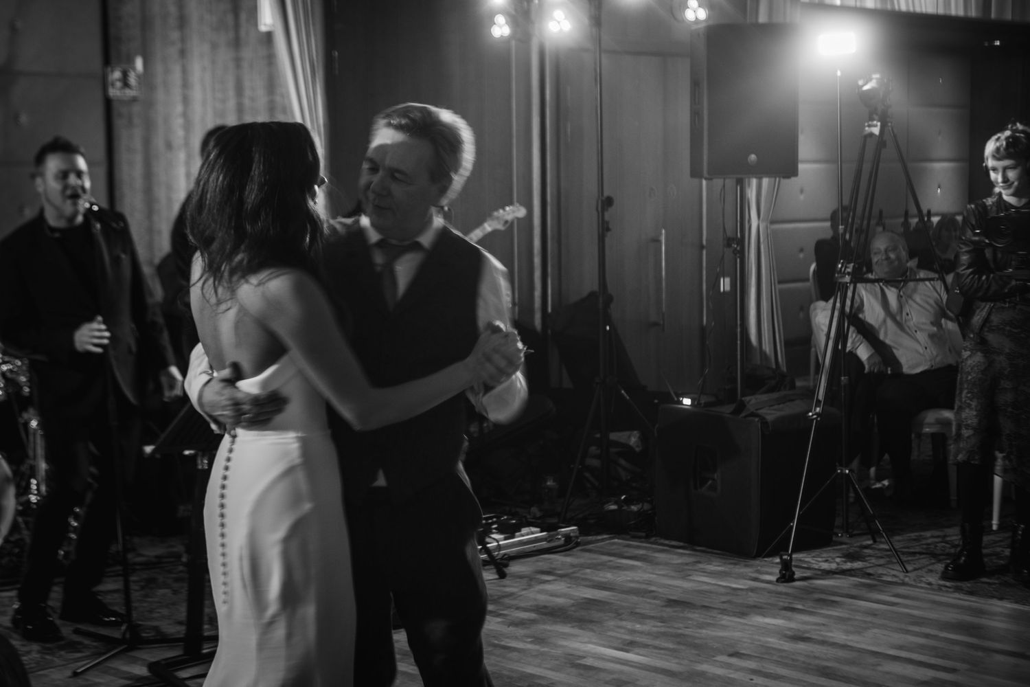 A couple shares their first dance at a dimly lit wedding reception while guests watch from the sidelines.
