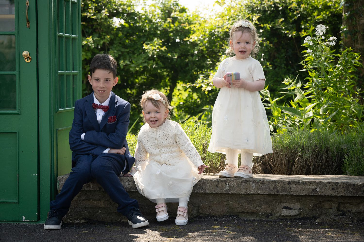 Three children in formal attire sit together on a stone wall near a green door.