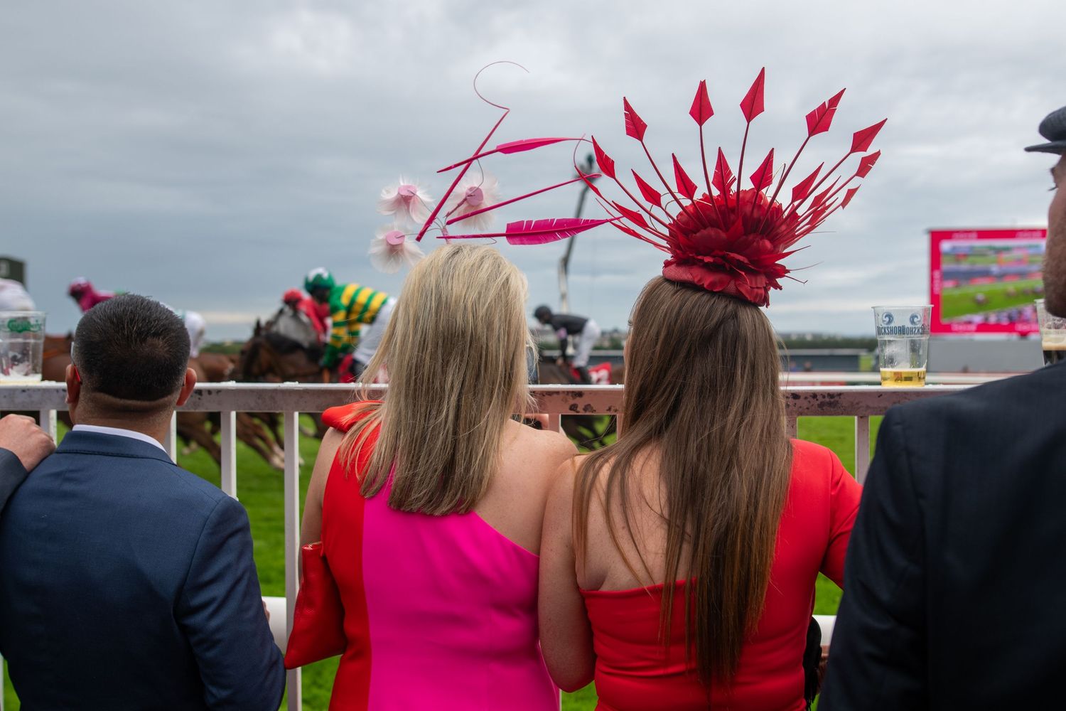 Spectators watching an outdoor event with vibrant red fascinators visible against cloudy sky.
