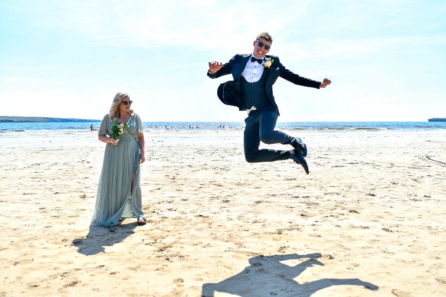 Playful beach wedding photo with person jumping while another stands on sandy shore.