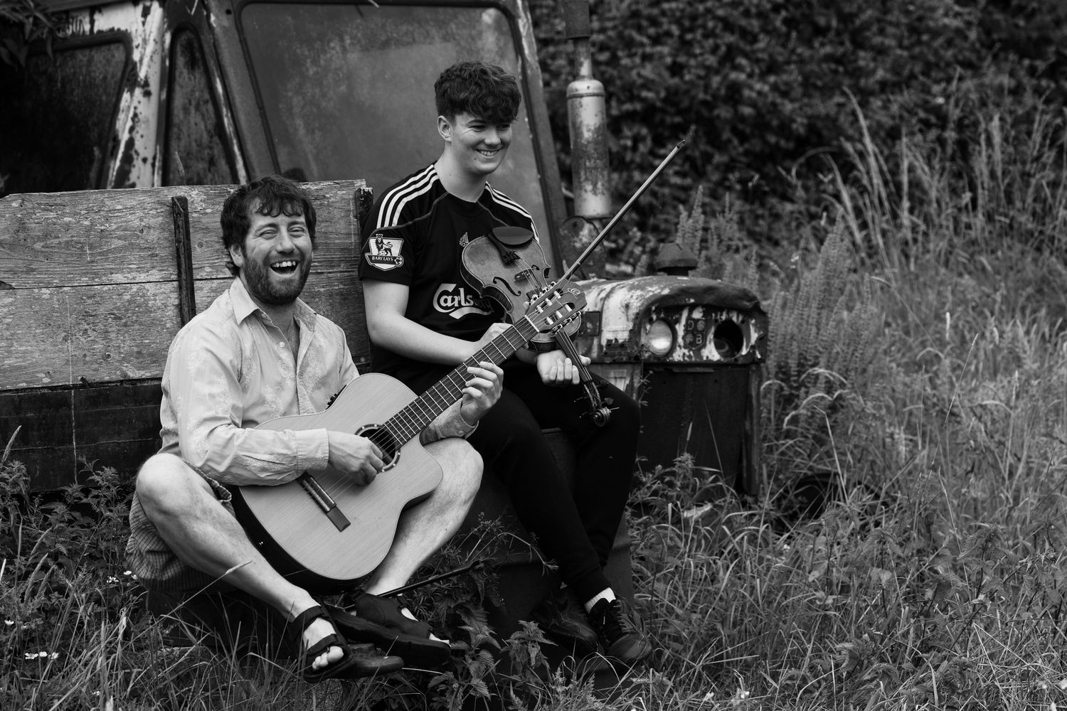 Two musicians play acoustic guitars while sitting in a grassy outdoor area.