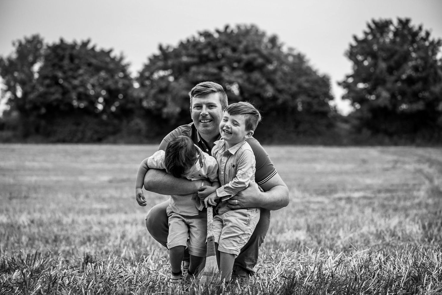 Black and white portrait of tender moment captured in an open field with trees in background.