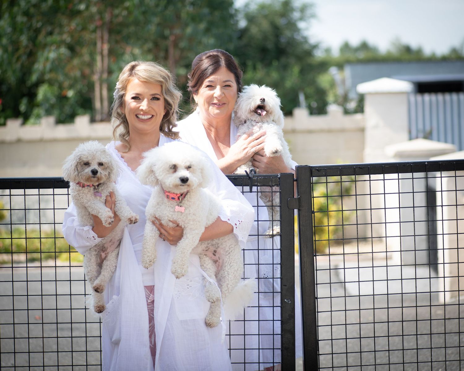 Two women in white dresses hold three small fluffy white dogs while standing by a fence.