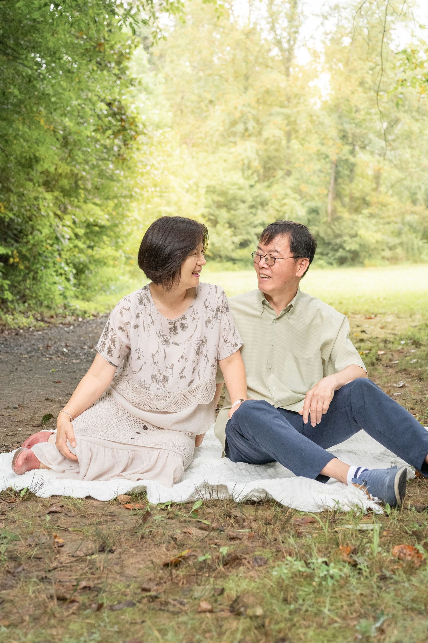 A couple shares a tender outdoor moment while lounging on a white blanket in a sunlit wooded area.
