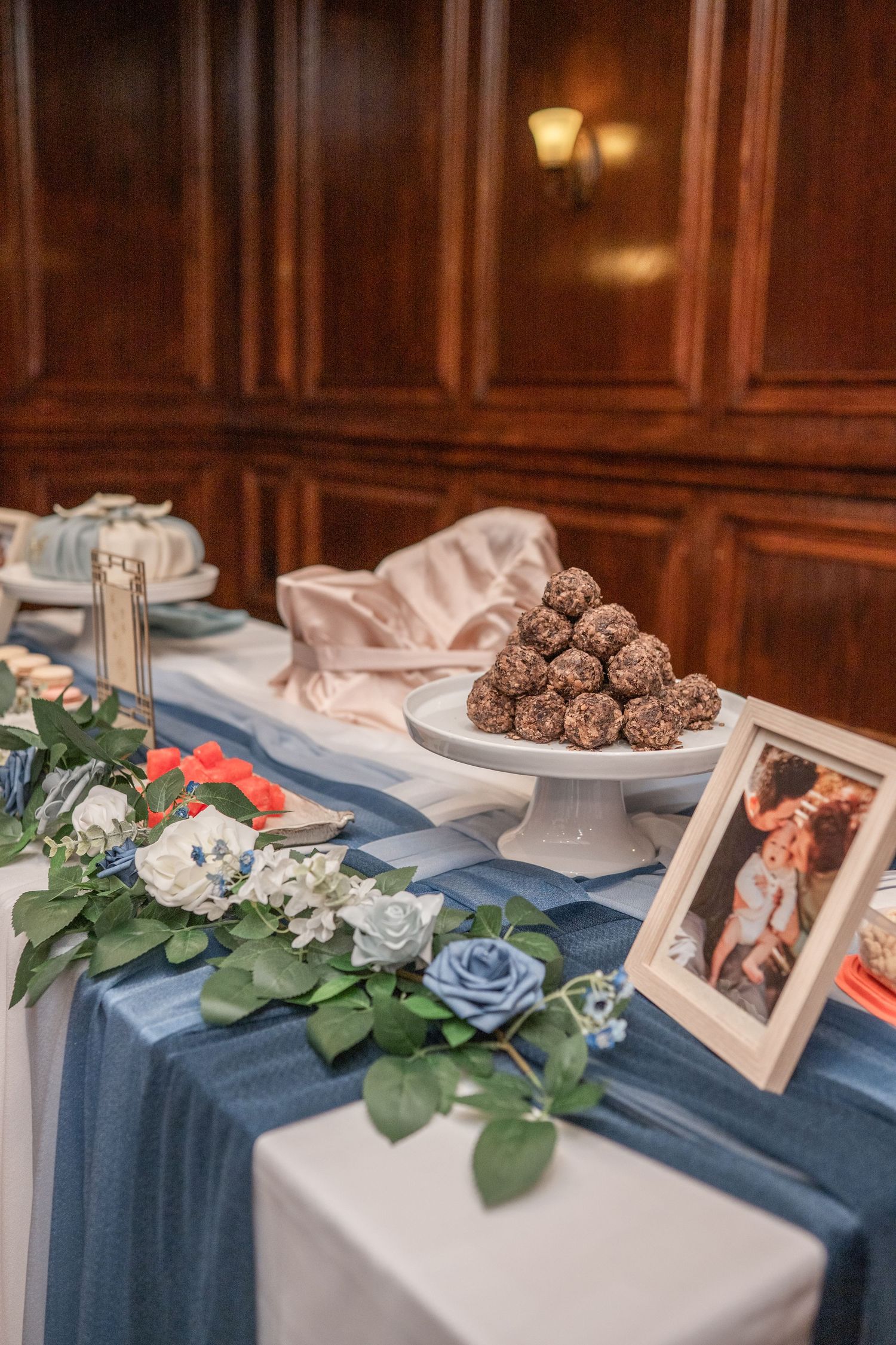Memorial table display with blue tablecloth decorated with framed photos, roses, and chocolate truffles at an event.